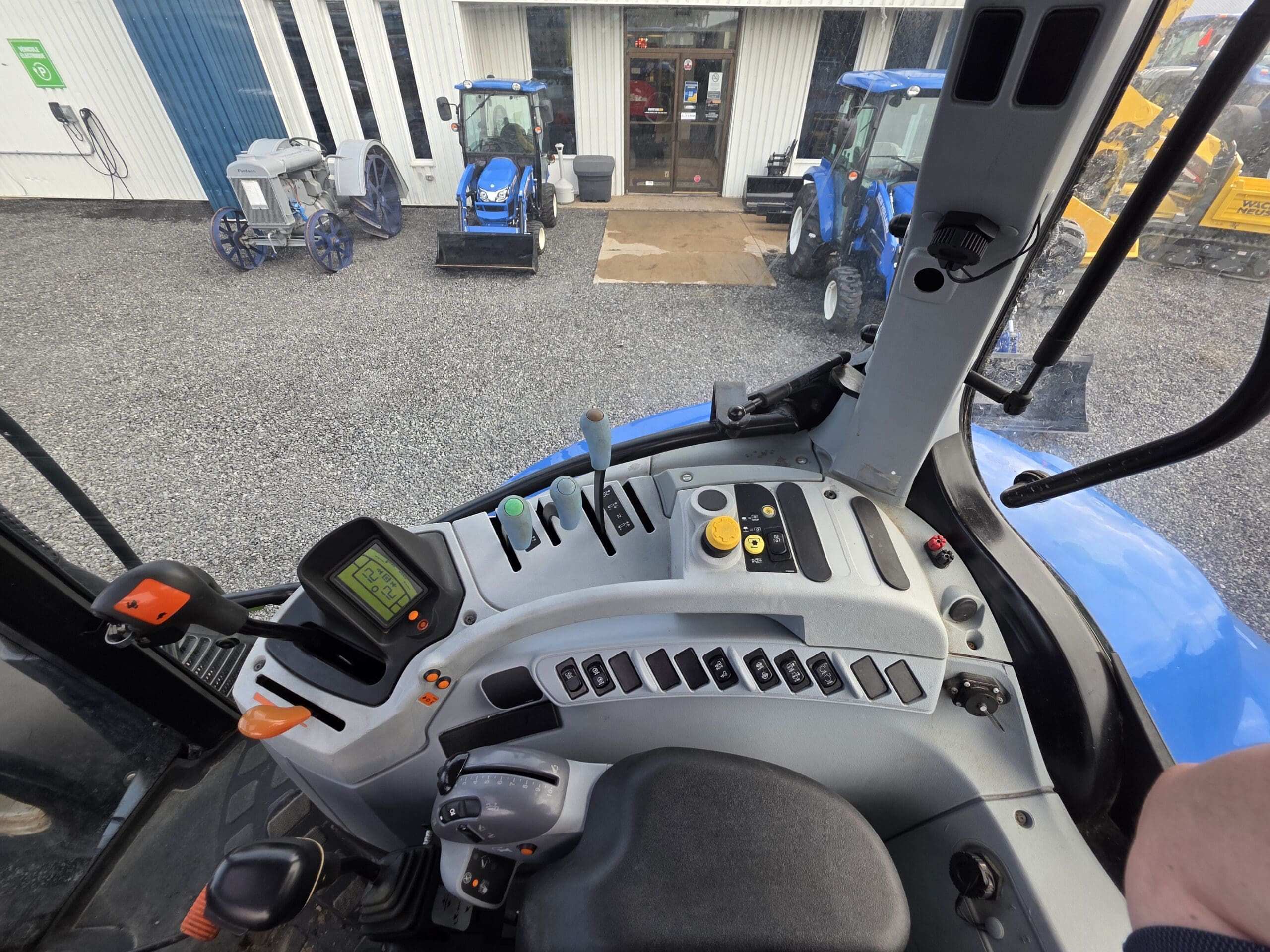 Vue de l'intérieur d'une cabine de tracteur montrant le panneau de commande, le volant et le siège. À l'extérieur, deux tracteurs bleus et d'autres équipements sont garés sur du gravier près de l'entrée d'un bâtiment.