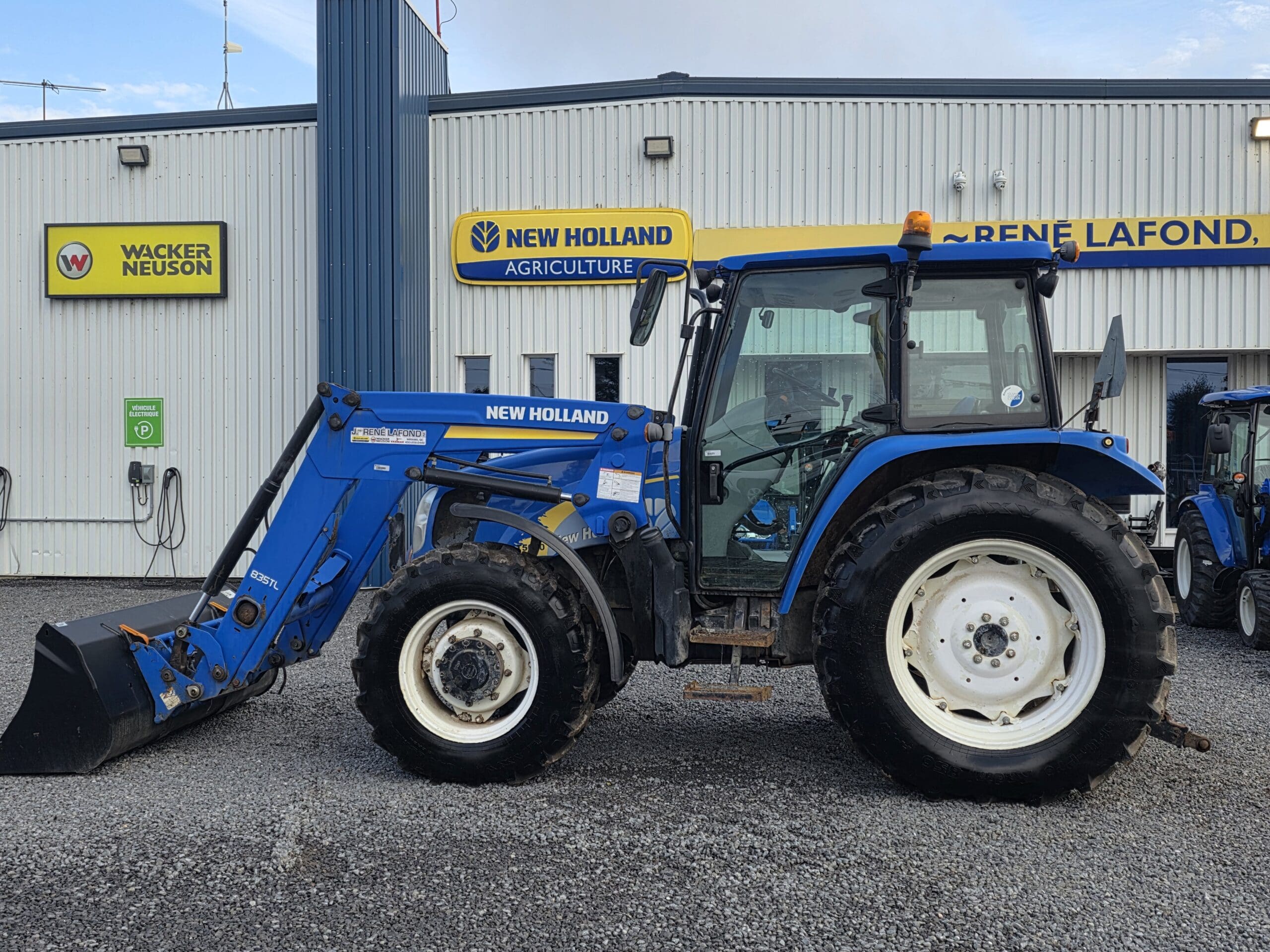 Un tracteur New Holland bleu équipé d'un chargeur frontal est garé sur du gravier devant un bâtiment portant des enseignes New Holland Agriculture et Wacker Neuson.