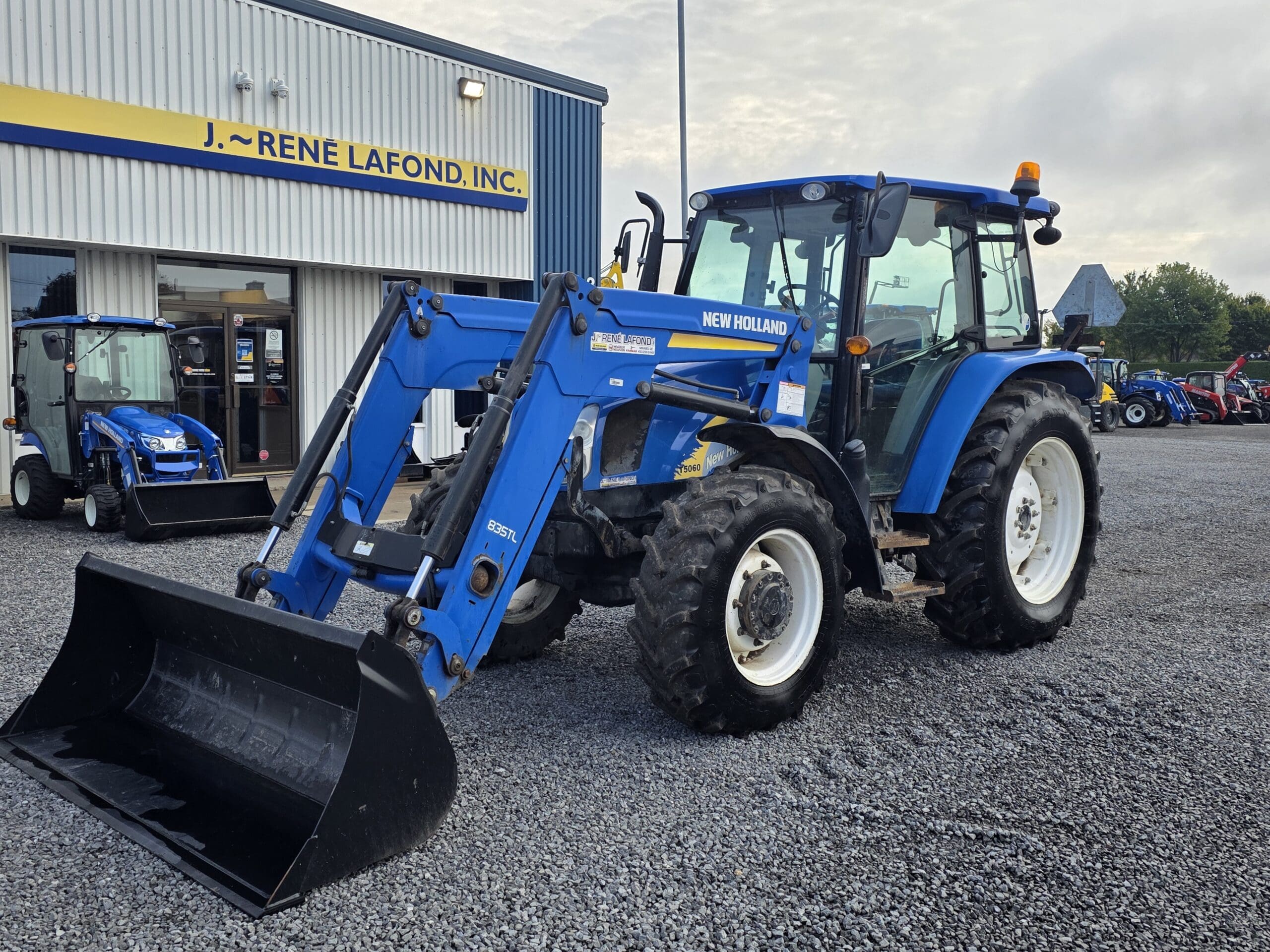 Un tracteur New Holland bleu équipé d'un chargeur frontal est garé sur du gravier à l'extérieur d'un bâtiment portant l'inscription 