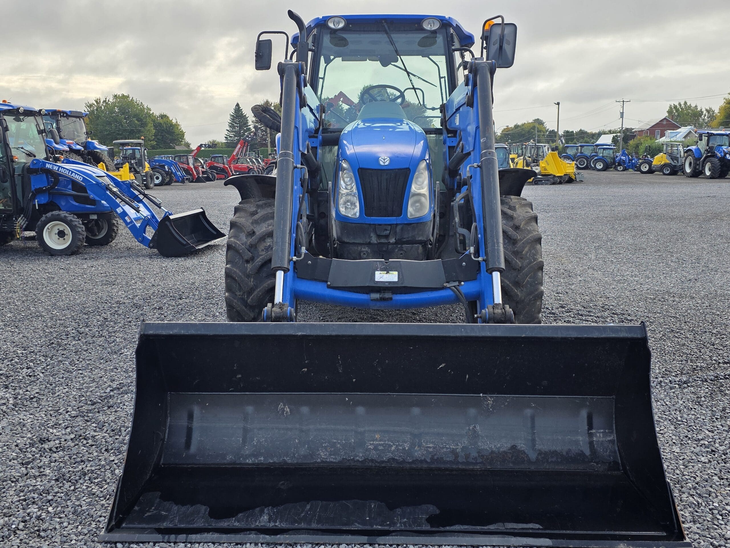 Un tracteur bleu équipé d'un chargeur frontal est garé sur un terrain en gravier, face à la caméra. D'autres tracteurs et équipements sont visibles à l'arrière-plan sous un ciel nuageux.