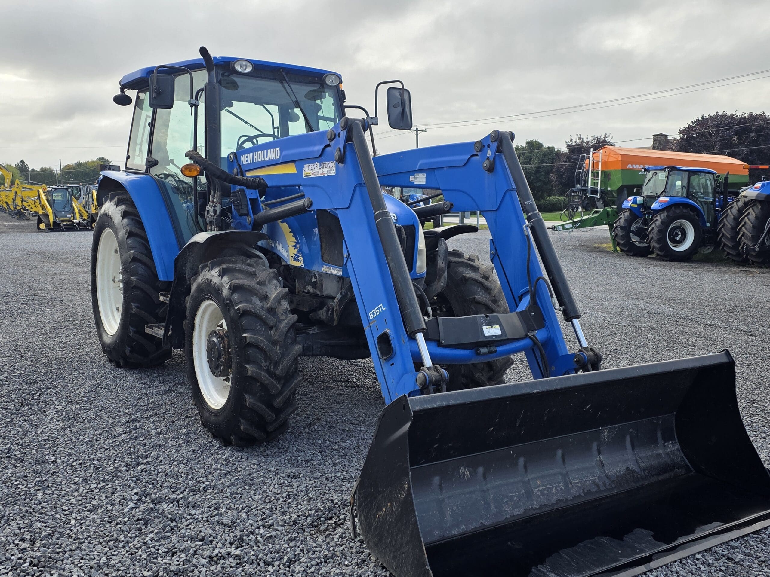 Un tracteur New Holland bleu équipé d'un chargeur frontal est garé sur un terrain en gravier. D'autres tracteurs sont visibles en arrière-plan sous un ciel nuageux.