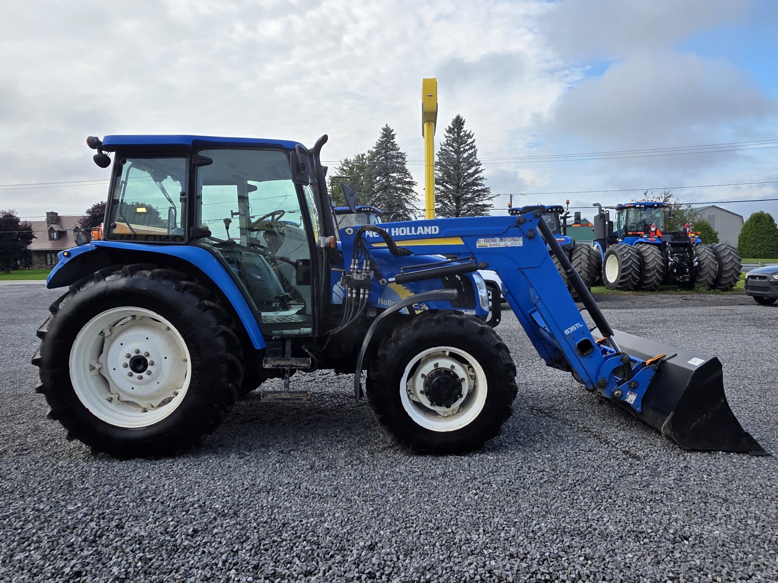 Un tracteur New Holland bleu équipé d'un chargeur frontal est garé sur un terrain en gravier. Un autre grand tracteur et des arbres sont visibles à l'arrière-plan.
