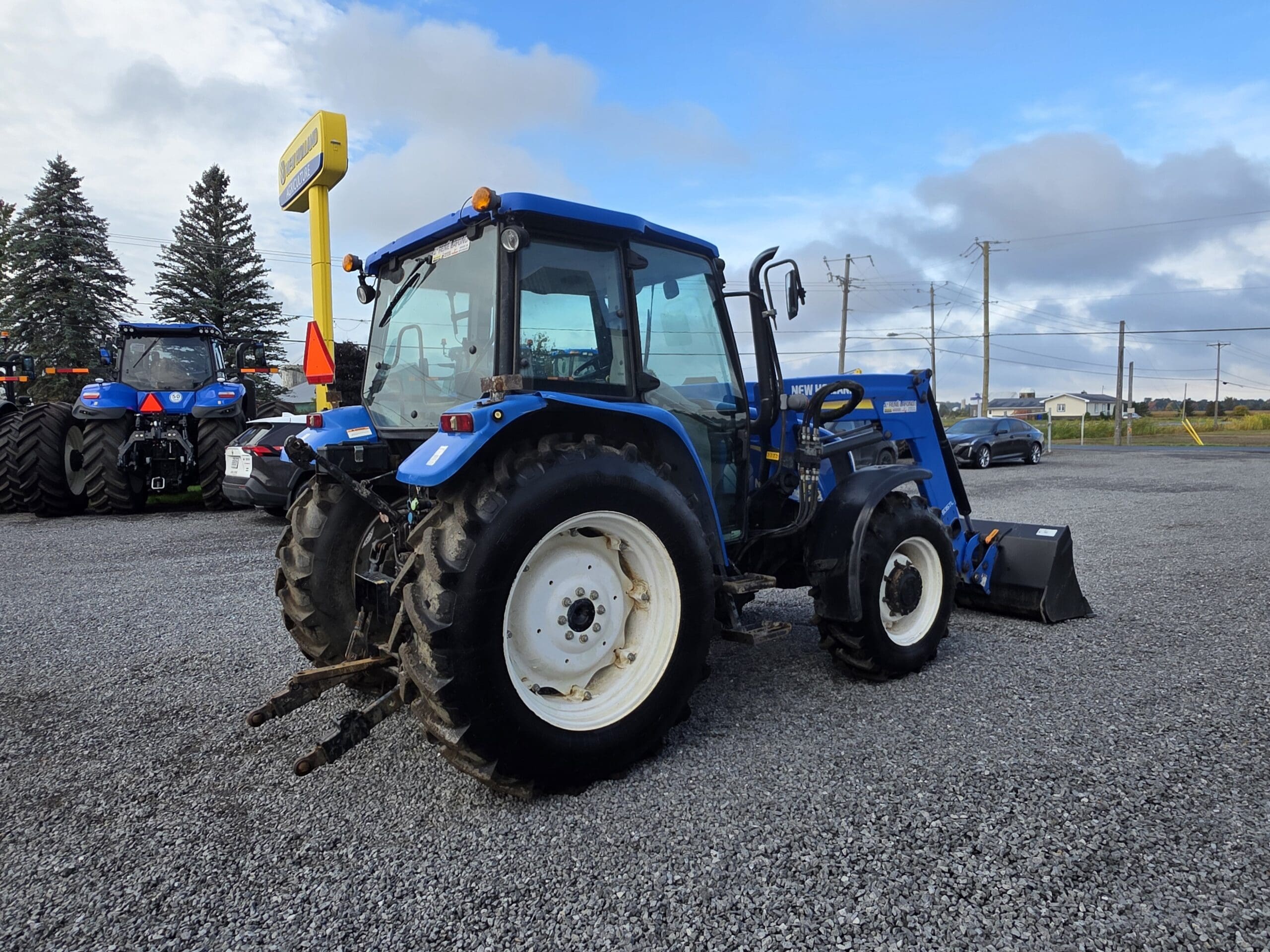 Un tracteur agricole New Holland bleu avec un chargeur frontal garé sur un terrain en gravier, avec un autre tracteur, des voitures garées et des arbres en arrière-plan sous un ciel partiellement nuageux.