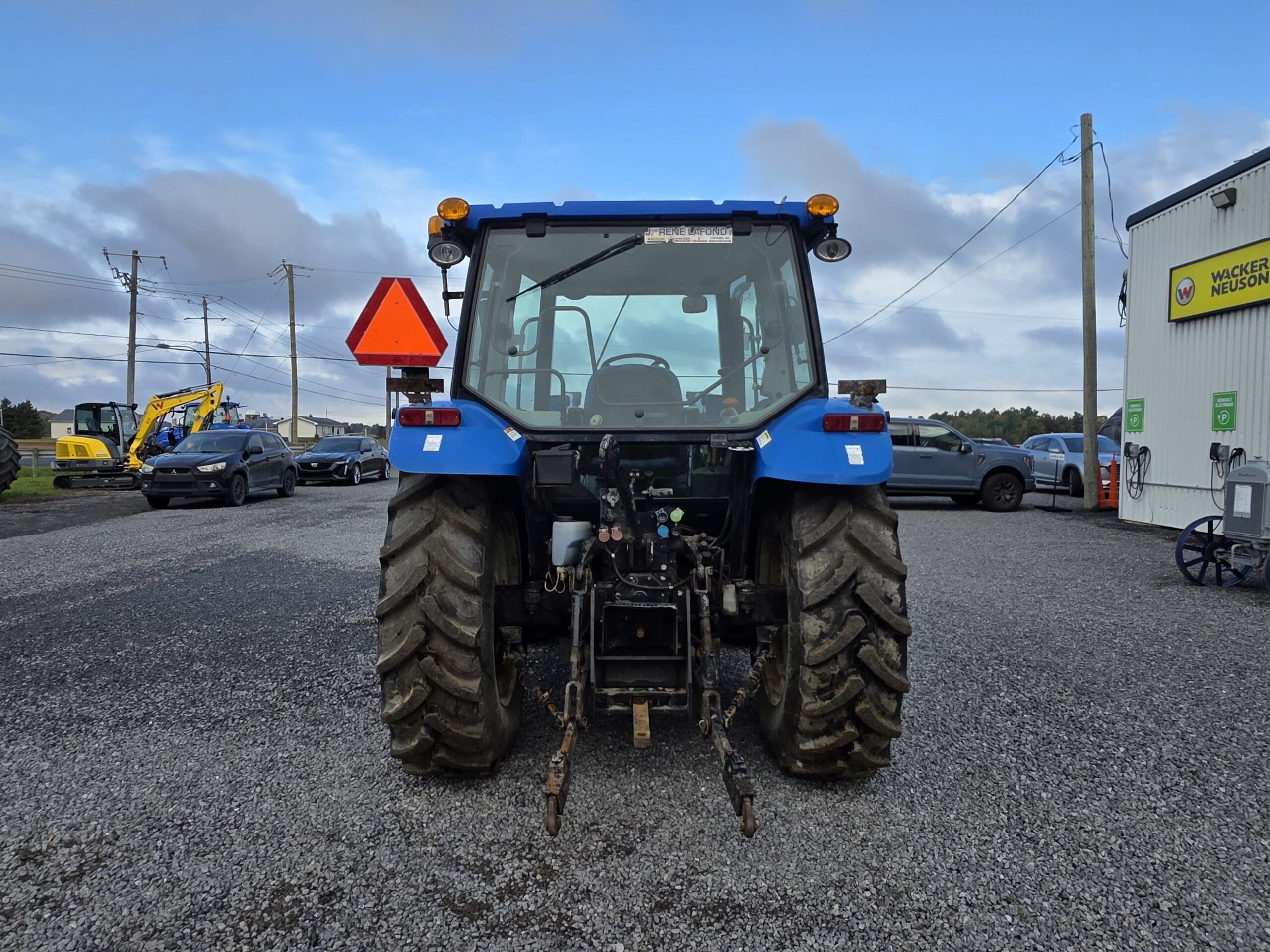 Un tracteur bleu est garé sur du gravier, vu de l'arrière. Il est entouré de voitures et d'engins de chantier sous un ciel partiellement nuageux.