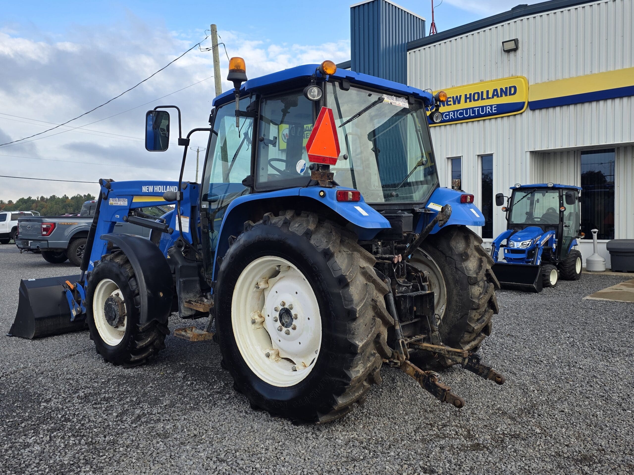 Un tracteur bleu New Holland est garé sur du gravier à l'extérieur d'une concession New Holland Agriculture. Un autre tracteur bleu plus petit est visible à l'arrière-plan, près du bâtiment.