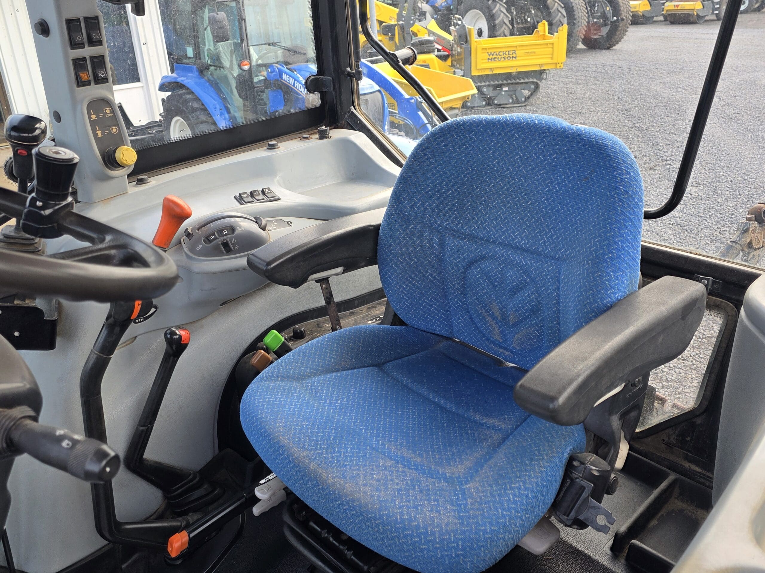 Intérieur d'une cabine de tracteur montrant un siège de conducteur à coussin bleu avec des accoudoirs, un volant, des leviers de commande et des panneaux de commande. La vue donne sur d'autres tracteurs et machines garés à l'extérieur.