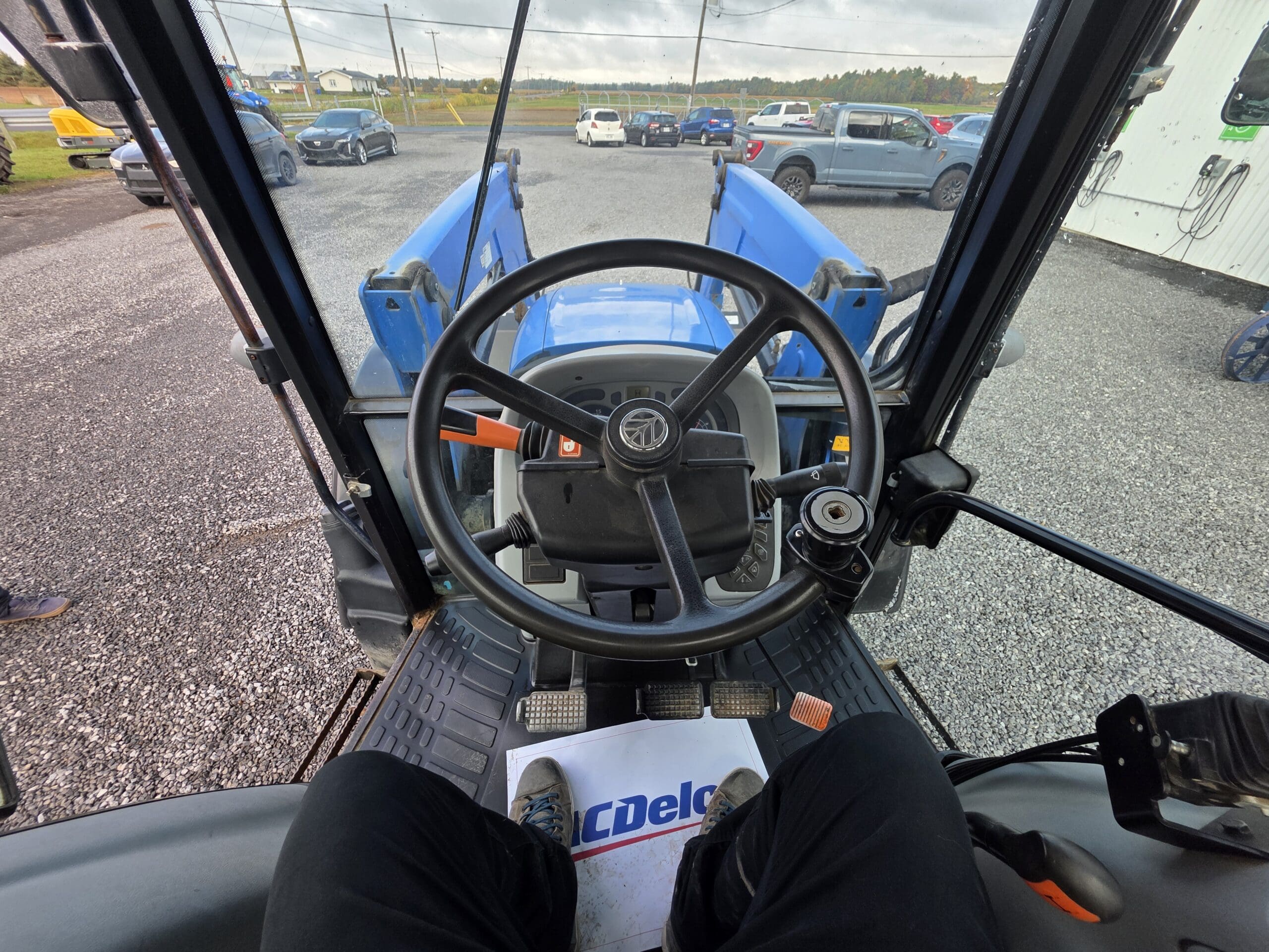 Vue de l'intérieur d'une cabine de tracteur, montrant le volant, le tableau de bord, les pédales et les jambes du conducteur. À l'extérieur, on voit des voitures garées et une route en arrière-plan sous un ciel nuageux.