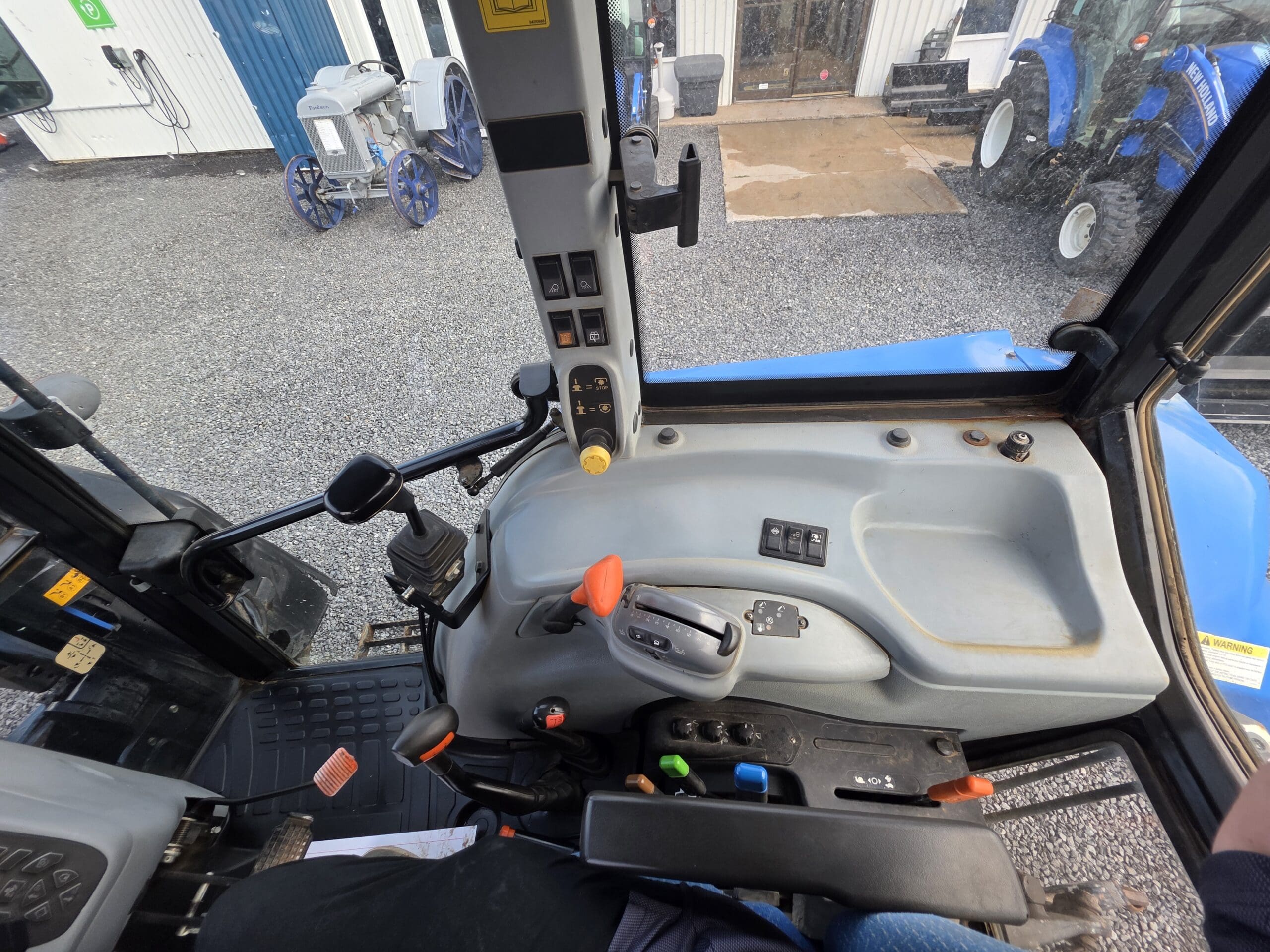 Vue de l'intérieur de la cabine d'un tracteur bleu montrant le panneau de commande, le volant, les leviers et divers boutons. Le sol à l'extérieur est en gravier, et d'autres tracteurs et équipements bleus sont visibles à l'arrière-plan.