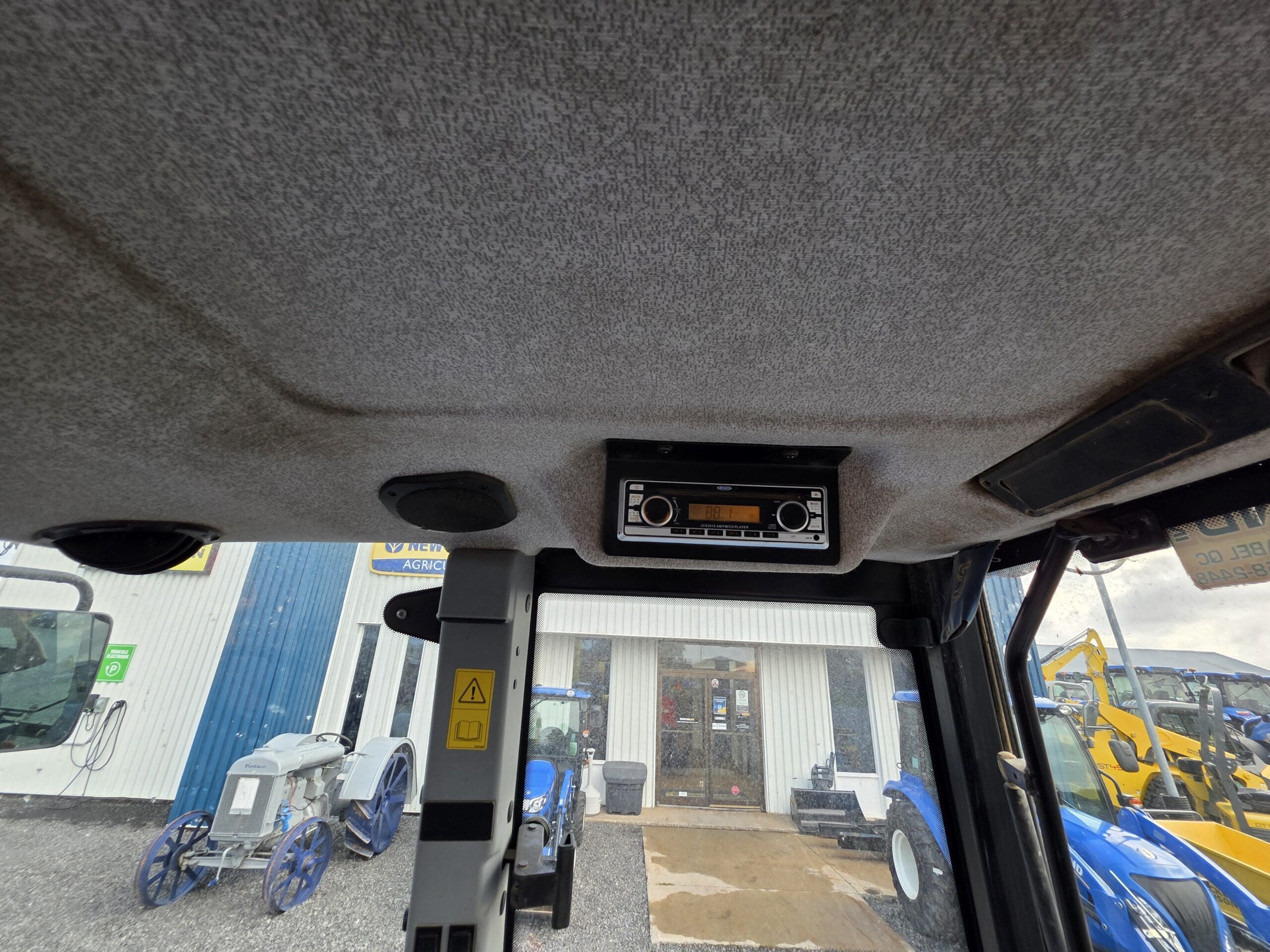 Vue de l'intérieur d'une cabine de tracteur regardant vers le haut une radio montée au plafond. À l'extérieur du pare-brise, on aperçoit des tracteurs bleus et un bâtiment avec des portes vitrées et du matériel agricole.