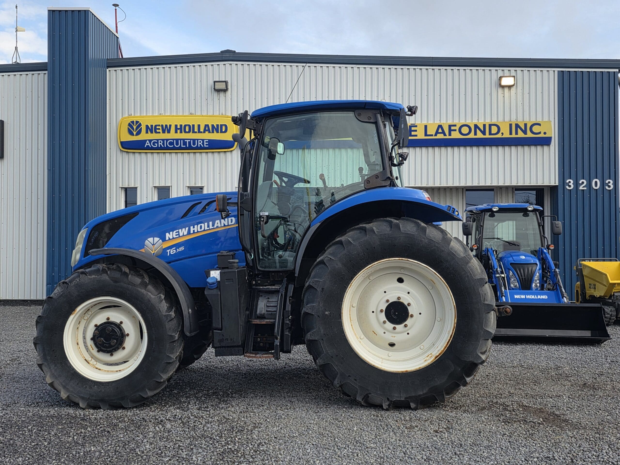 Un tracteur bleu New Holland T6.180 est garé devant un bâtiment portant les inscriptions 