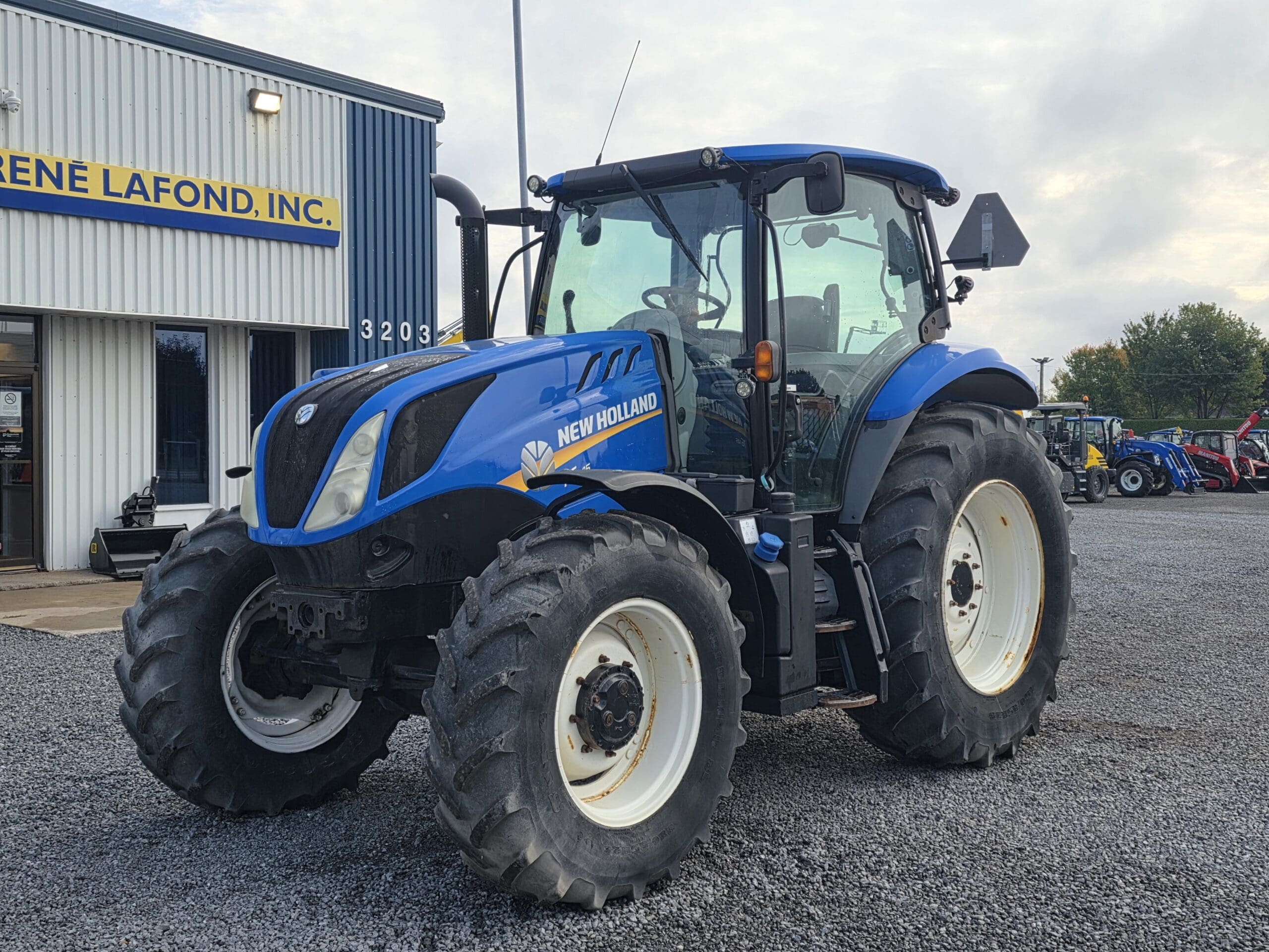 Un tracteur New Holland bleu est garé sur du gravier à l'extérieur d'un bâtiment portant un panneau indiquant 