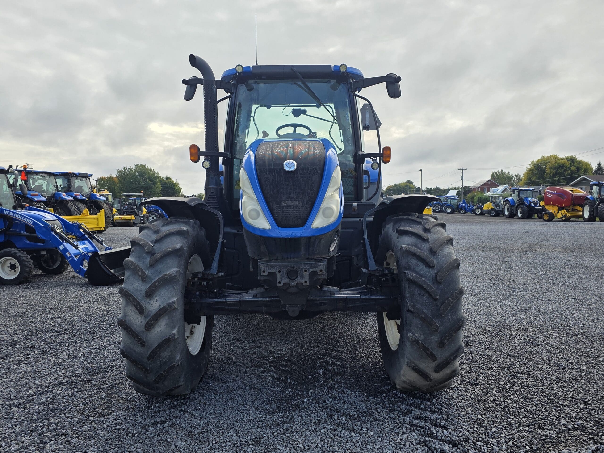 Un tracteur bleu et noir est garé sur du gravier, face à la caméra. D'autres machines agricoles et tracteurs sont visibles à l'arrière-plan sous un ciel nuageux.
