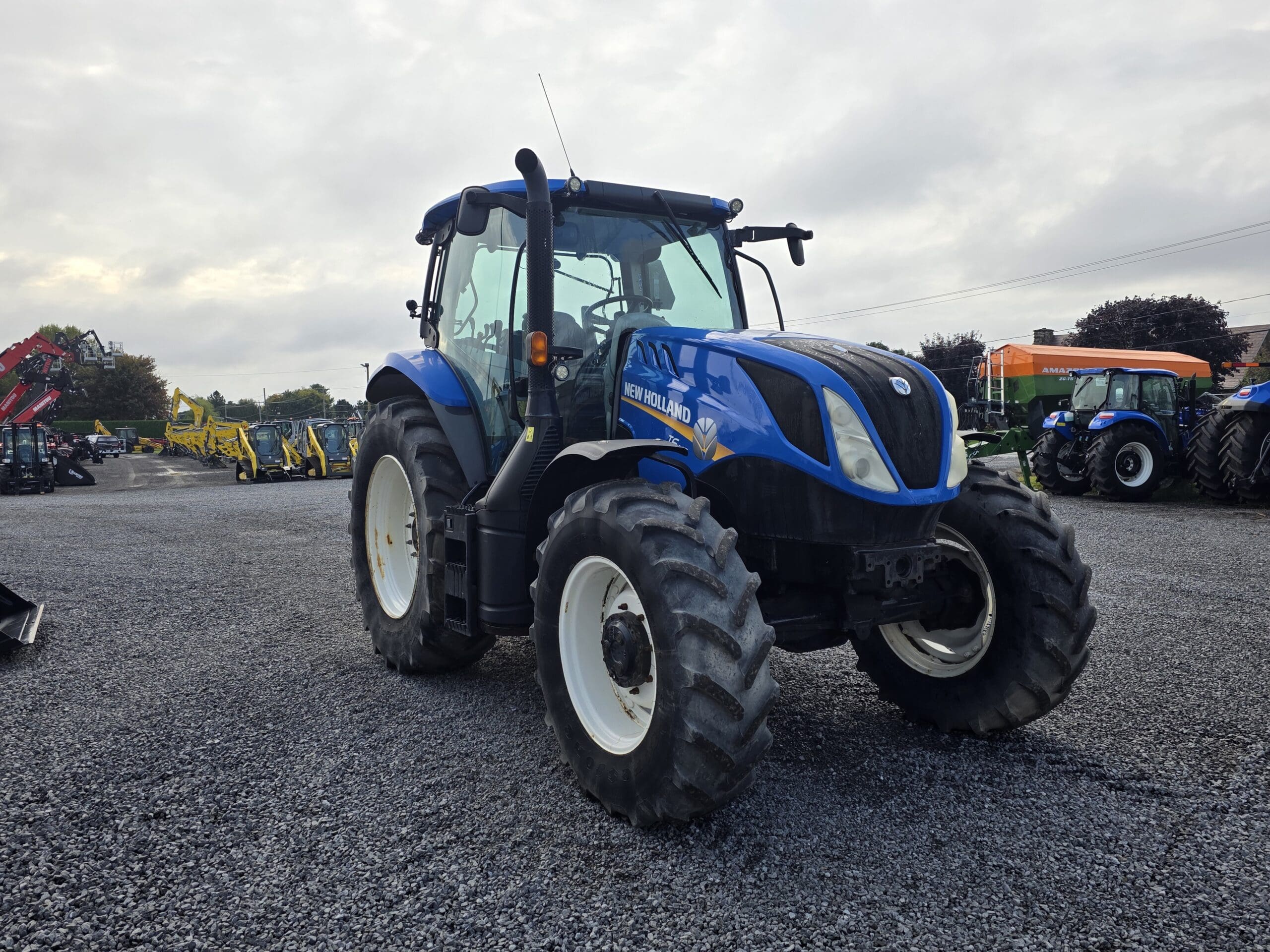 Un tracteur New Holland bleu est garé sur un terrain en gravier, entouré d'autres équipements agricoles et de tracteurs, sous un ciel nuageux.