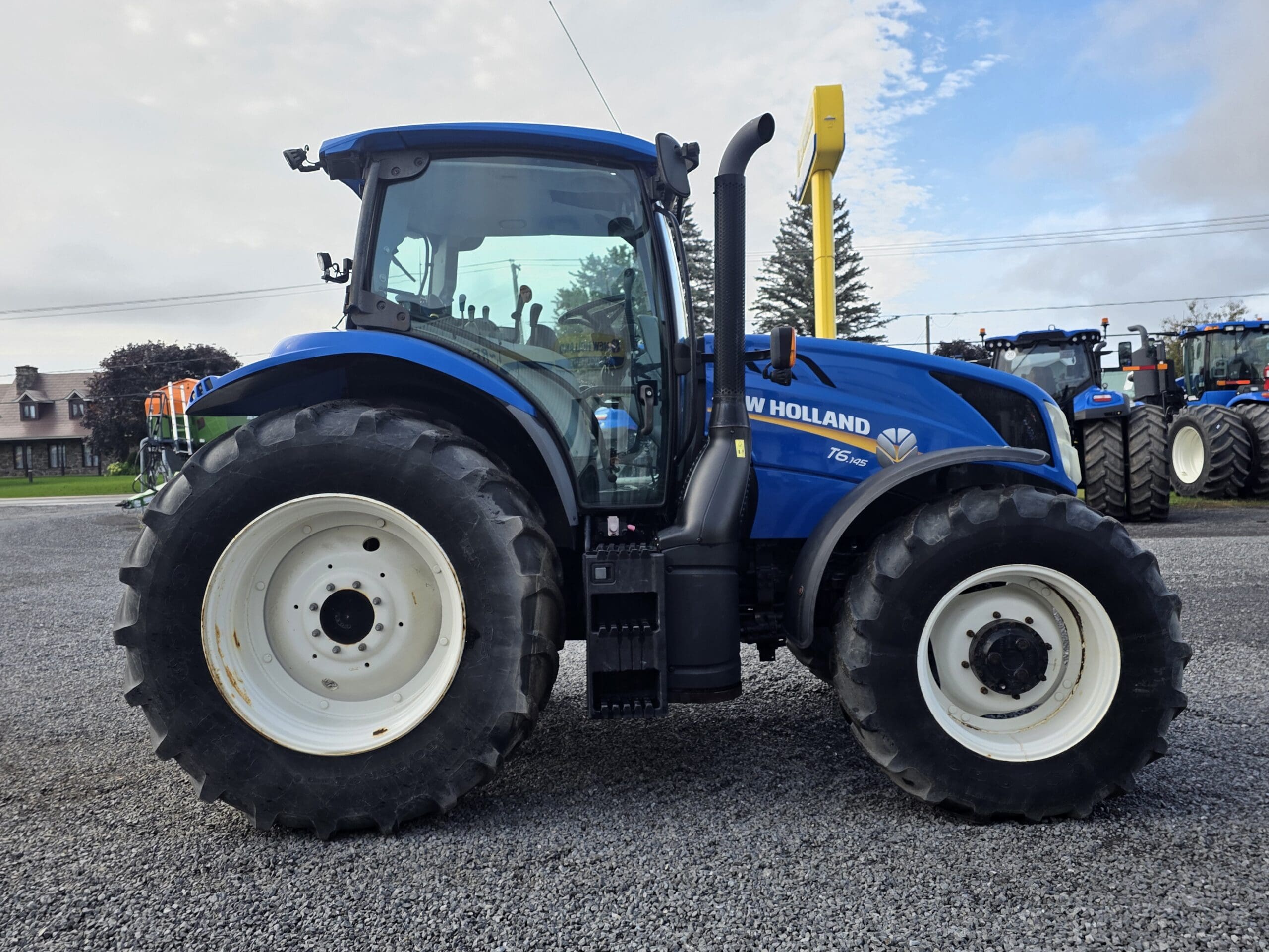 Un tracteur New Holland T5.95 bleu est garé sur un terrain en gravier, avec de gros pneus, des jantes blanches et des fenêtres transparentes ; d'autres tracteurs et des arbres sont visibles à l'arrière-plan.