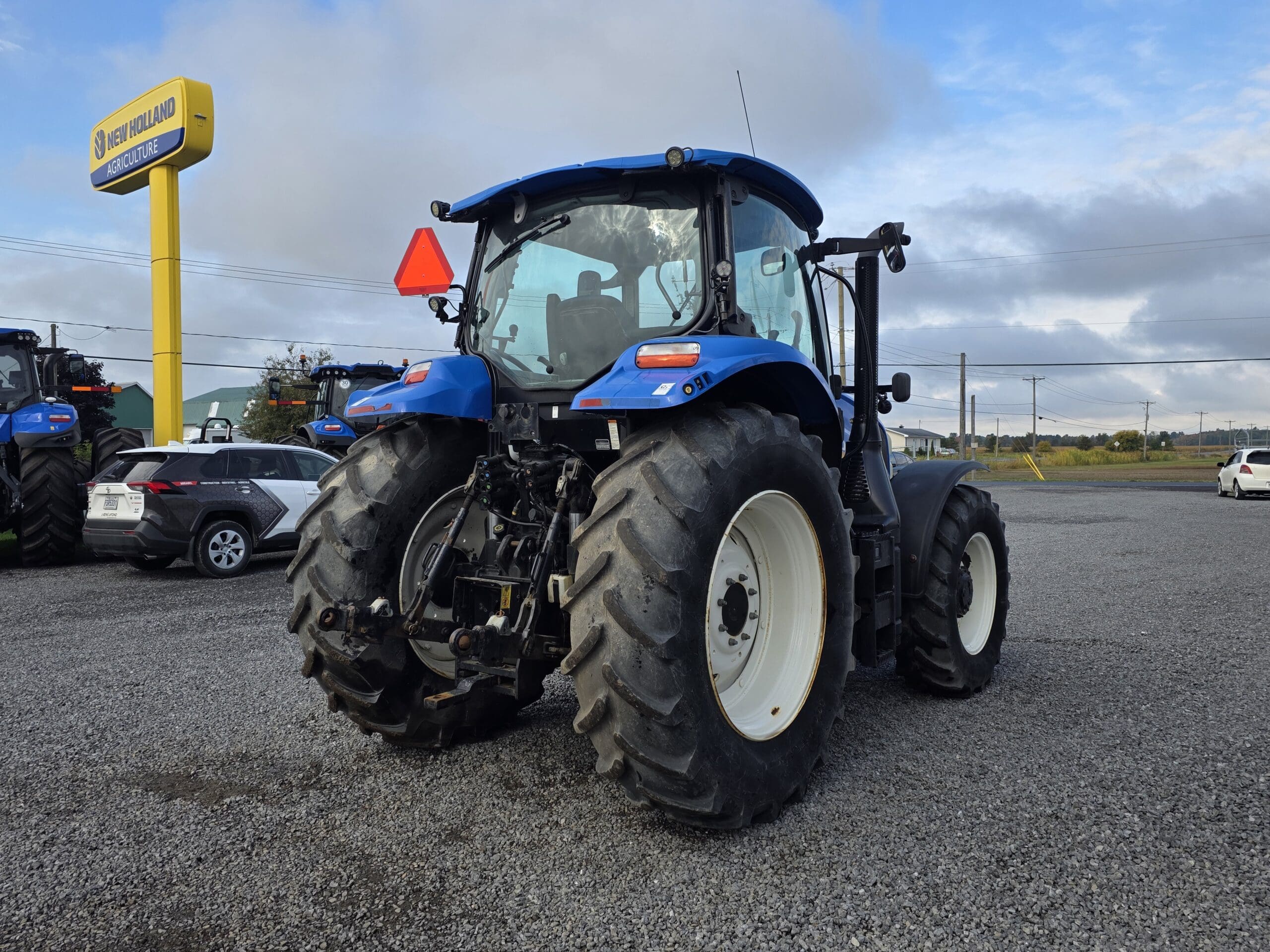 Un grand tracteur bleu avec de gros pneus arrière est garé sur un terrain en gravier près de l'enseigne d'un concessionnaire New Holland. D'autres véhicules et machines sont visibles à l'arrière-plan sous un ciel nuageux.