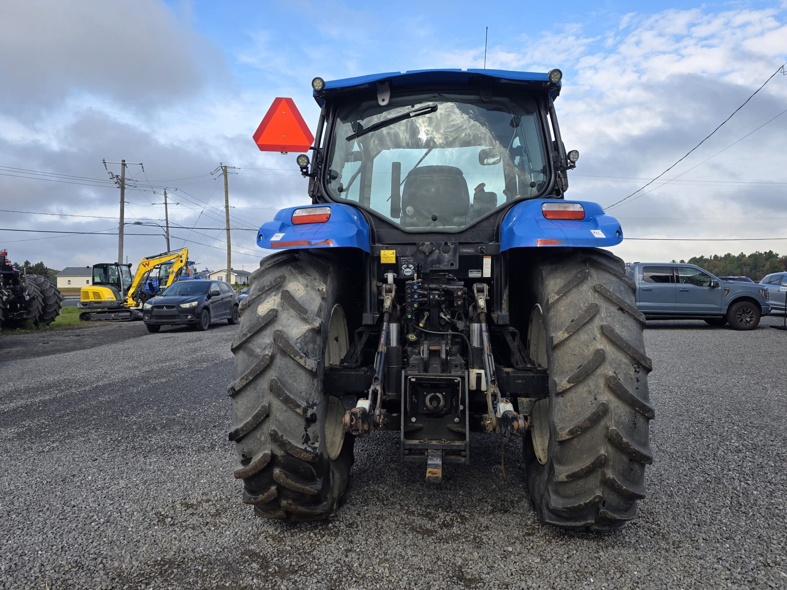Un tracteur bleu est garé sur un terrain en gravier, vu de l'arrière. Ses gros pneus sont boueux et un panneau d'avertissement triangulaire rouge se trouve à l'arrière. Des voitures et des engins de chantier sont visibles à l'arrière-plan.