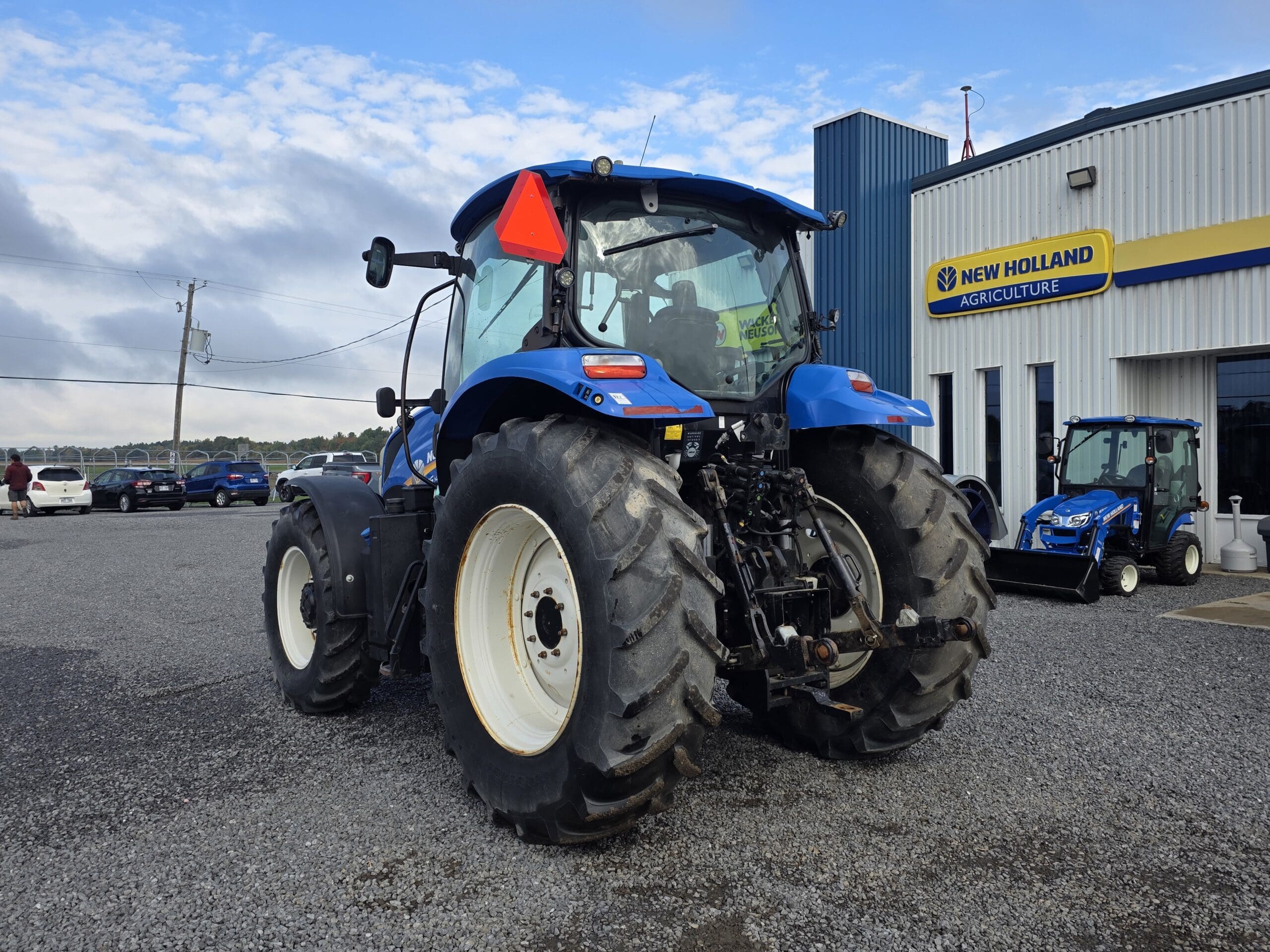 Un tracteur bleu est garé sur du gravier à l'extérieur d'une concession New Holland Agriculture, avec plusieurs voitures et un véhicule utilitaire bleu à l'arrière-plan, par une journée partiellement nuageuse.