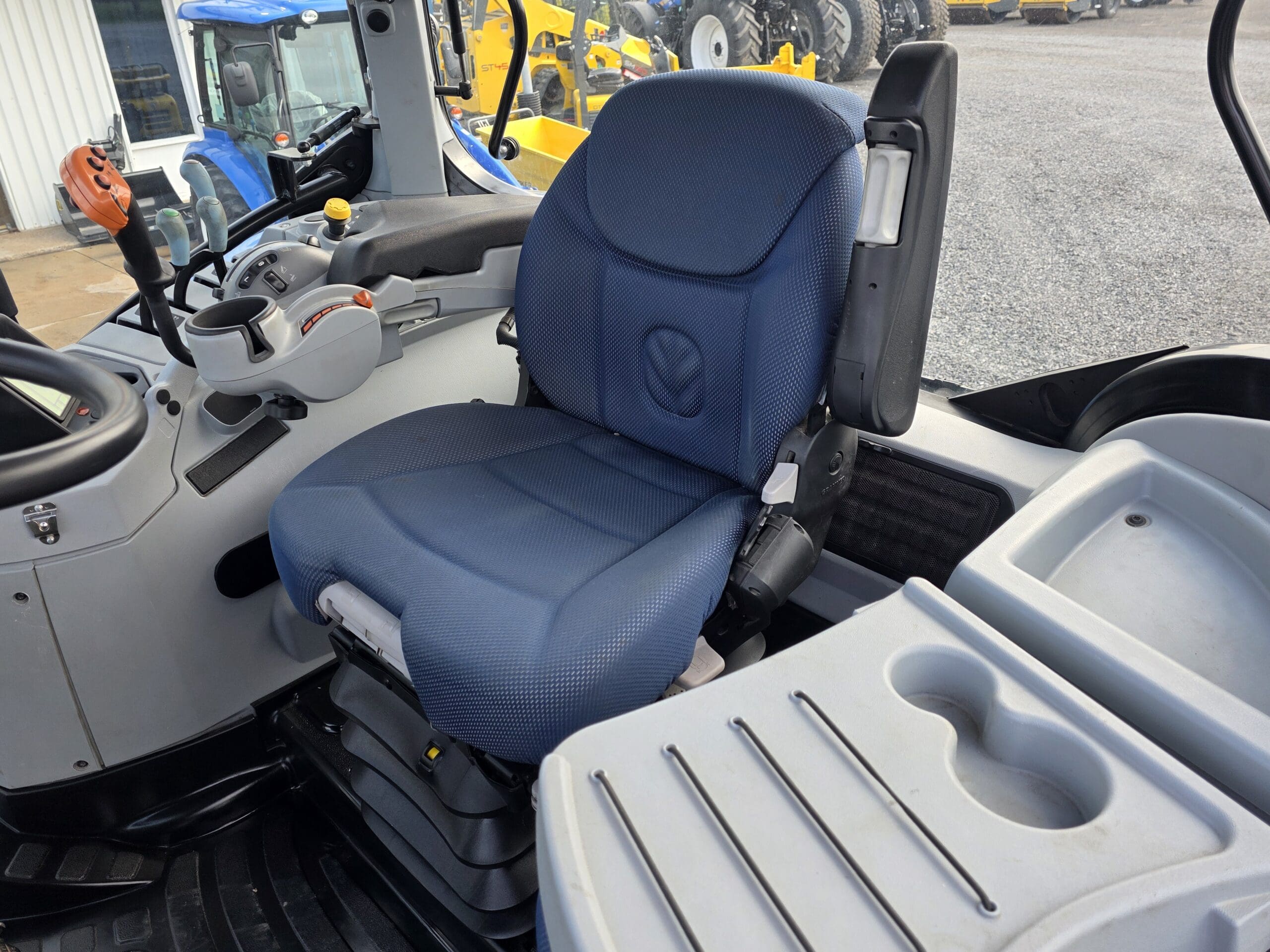 Gros plan sur le siège bleu du conducteur à l'intérieur d'une cabine de tracteur moderne, avec un panneau de commande, un accoudoir et des compartiments de rangement visibles à proximité. Les pneus et l'équipement du tracteur sont visibles à l'extérieur par la fenêtre.