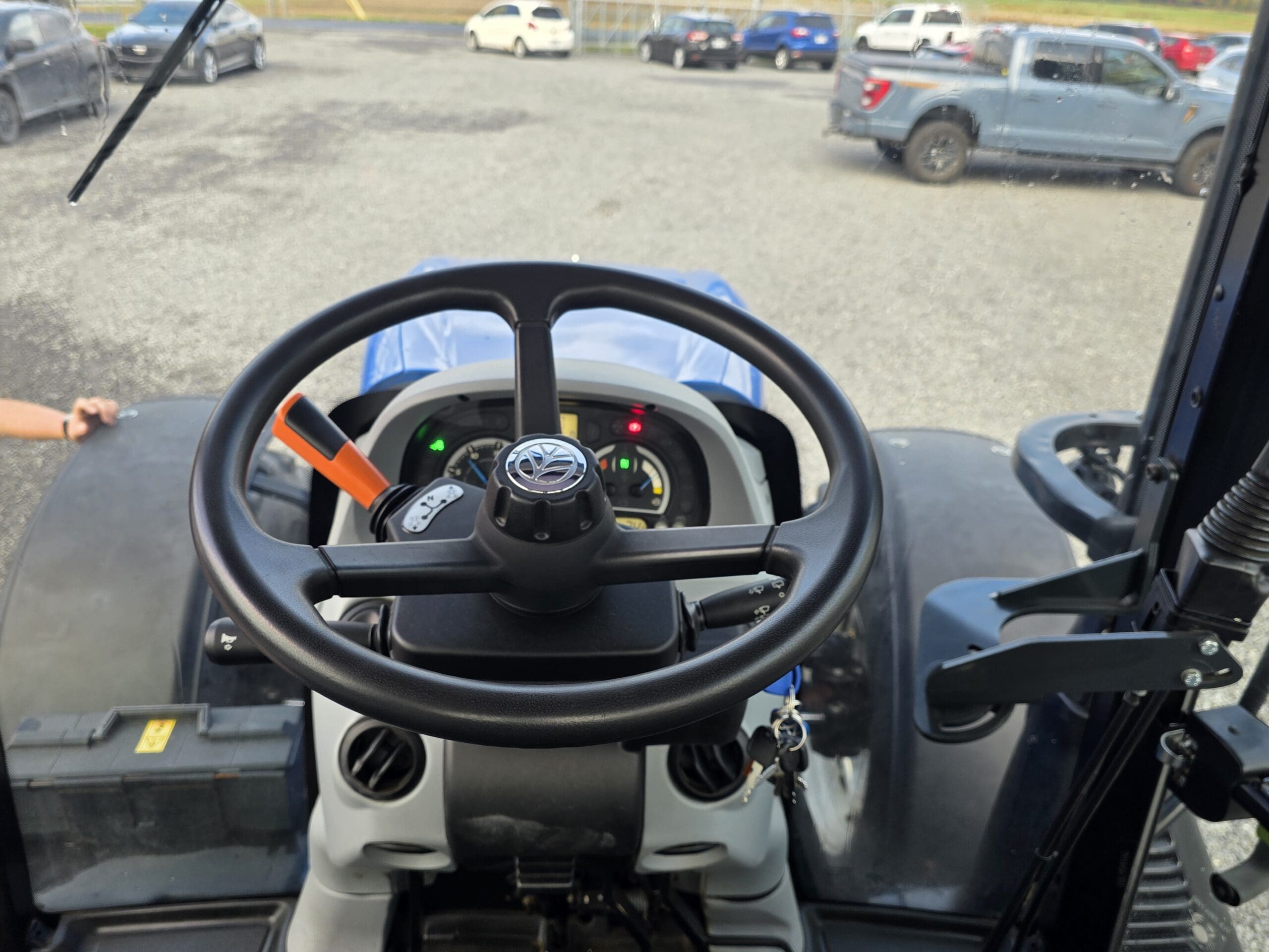 Vue du siège du conducteur d'un tracteur, montrant le volant, le tableau de bord avec des commandes éclairées, et une vue partielle du capot et du parking avec plusieurs voitures en arrière-plan.