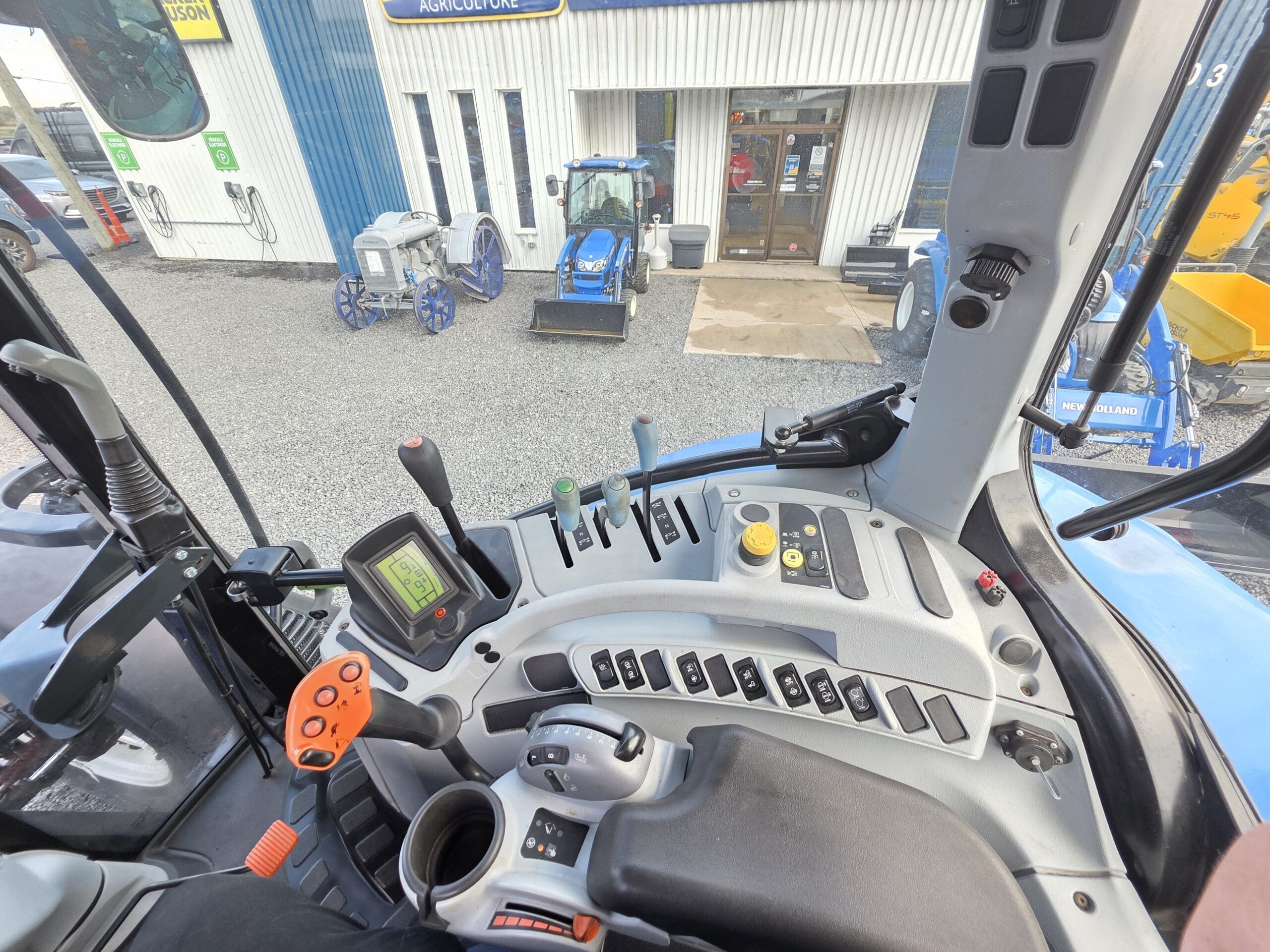 Vue de l'intérieur d'une cabine de tracteur moderne montrant le volant, le panneau de commande avec divers leviers et interrupteurs, et l'affichage numérique, en regardant vers les tracteurs et l'équipement garés devant un bâtiment.