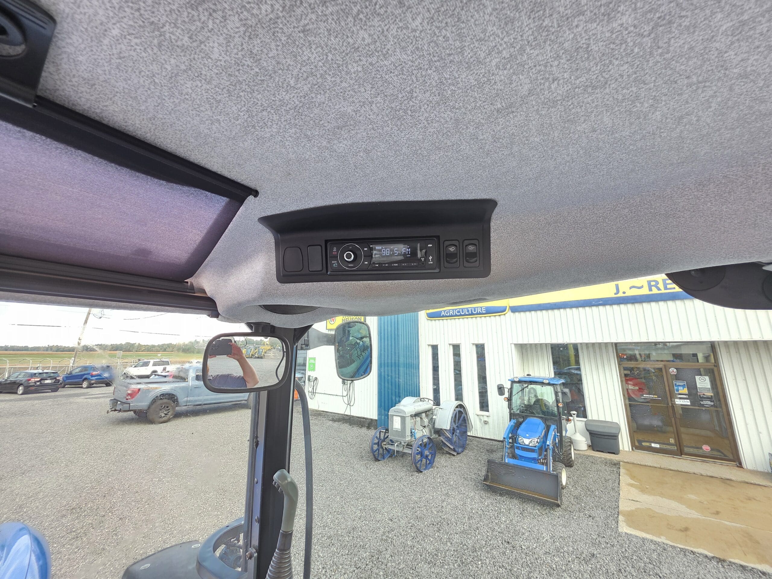 Vue intérieure d'une cabine de tracteur montrant un panneau de commande fixé au plafond. Par la fenêtre, plusieurs véhicules, du matériel et un tracteur bleu sont visibles à l'extérieur, devant un bâtiment portant une enseigne jaune et bleue.