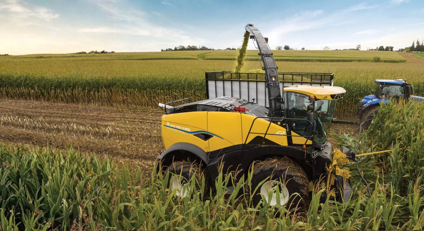 Une moissonneuse jaune de notre sélection de machinerie neuve et usagée coupe et transforme le maïs dans un grand champ. Les récoltes coupées sont chargées dans une remorque tirée par un tracteur bleu sous un ciel partiellement nuageux.