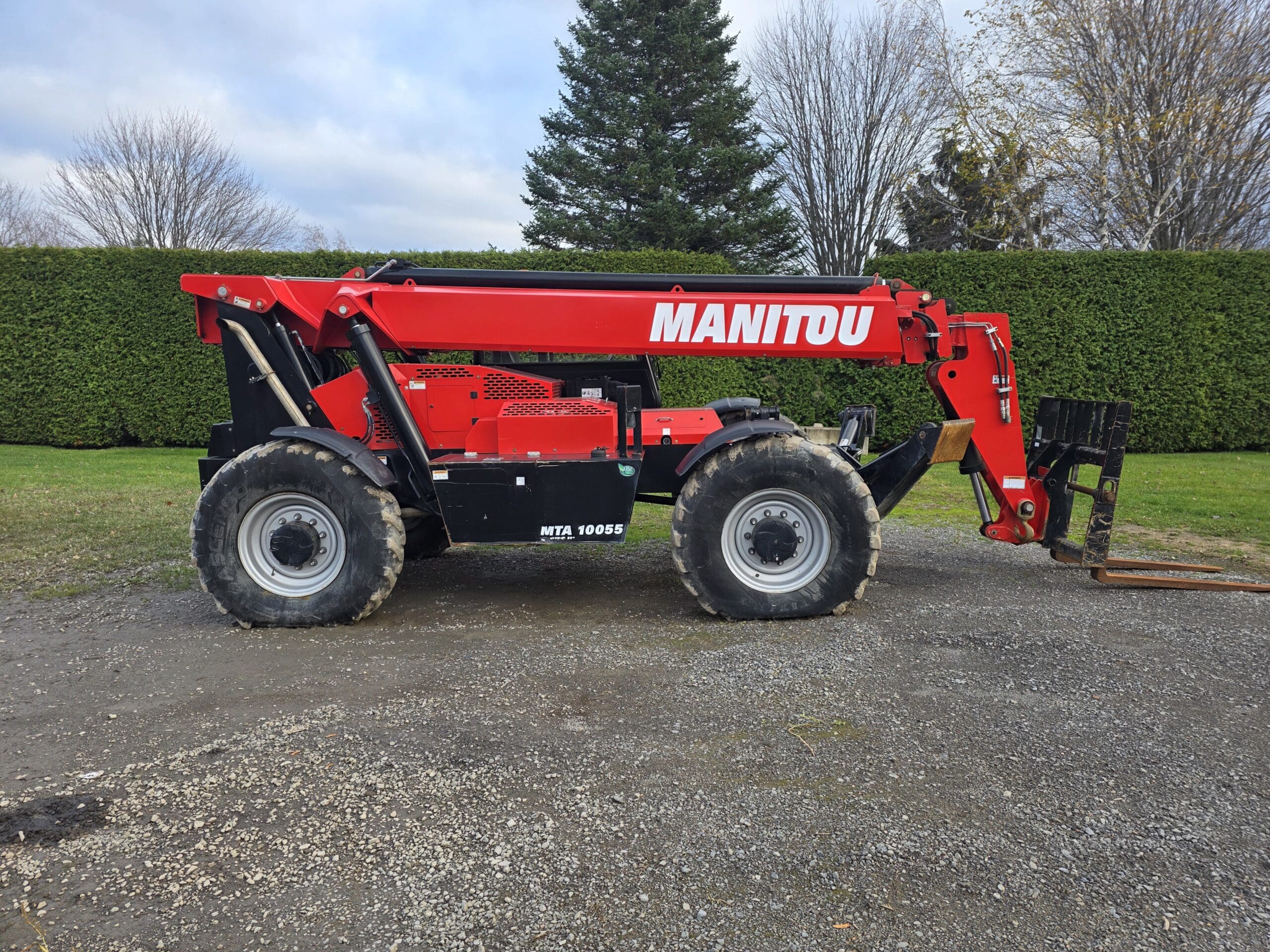Un chariot télescopique Manitou rouge à gros pneus est garé sur une aire de gravier, avec des haies vertes, de l'herbe et des arbres en arrière-plan, sous un ciel nuageux.