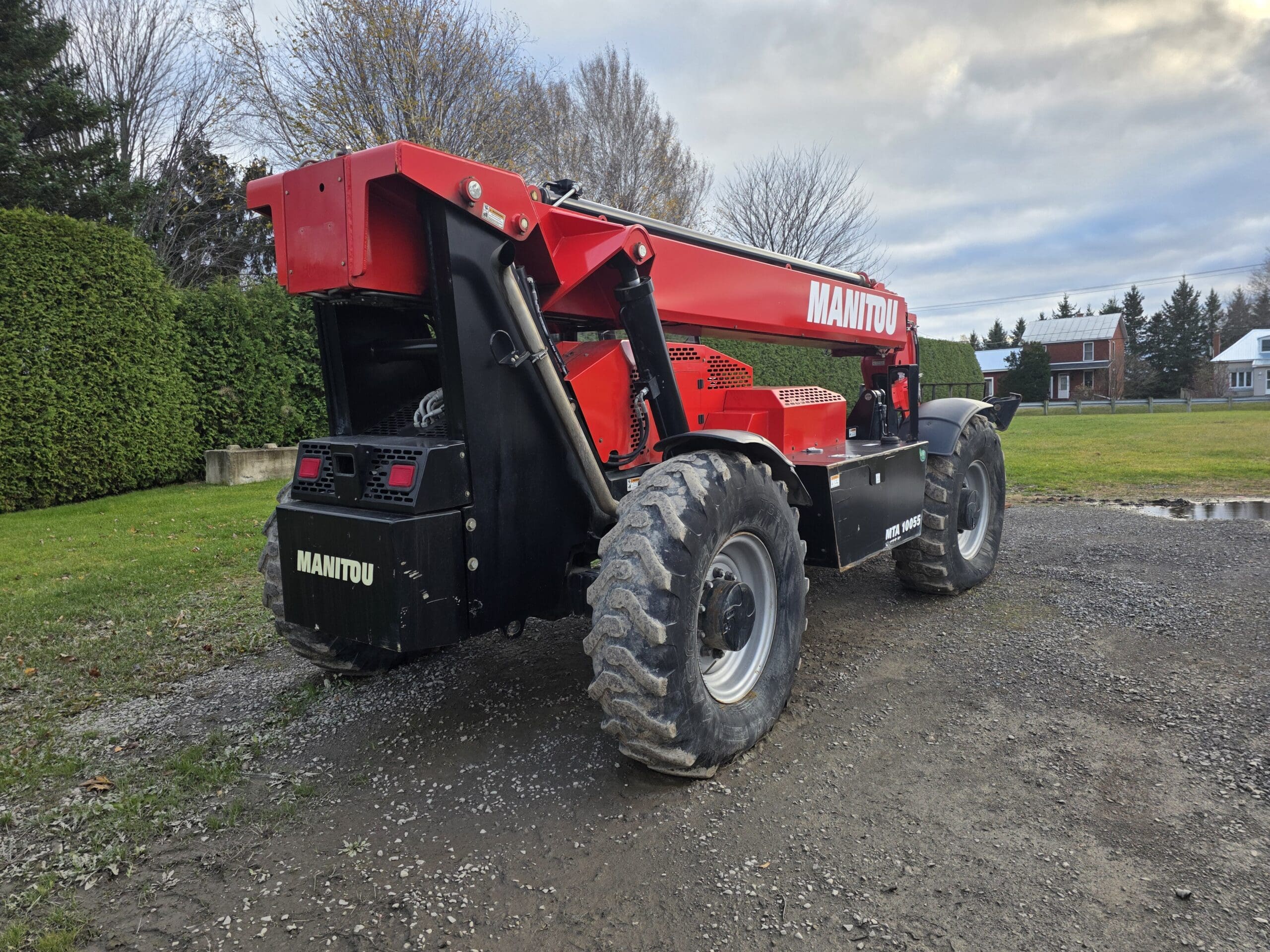 Un chariot télescopique Manitou rouge à gros pneus est garé sur une allée en gravier, entouré d'herbe verte et d'arbres, avec une maison et un ciel nuageux en arrière-plan.