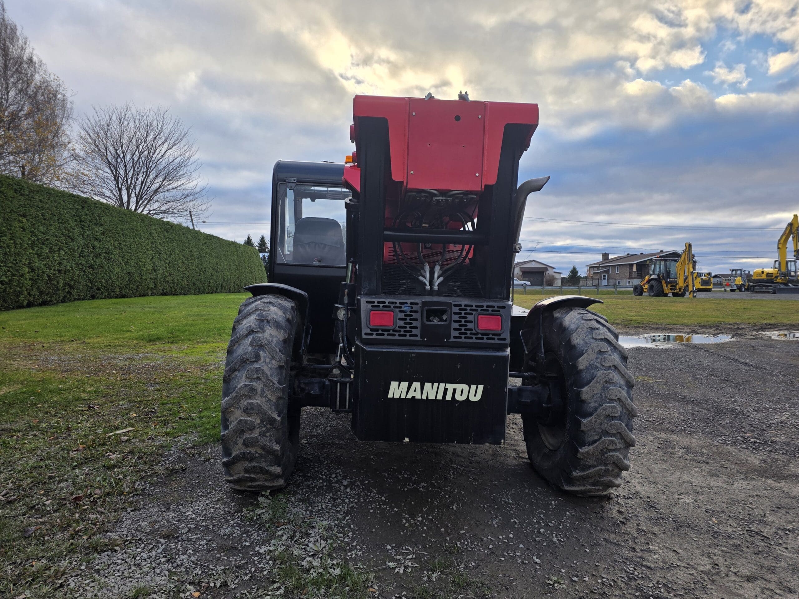 Vue arrière d'un grand tracteur MANITOU boueux garé sur un chemin de gravier, avec en arrière-plan de l'herbe verte, des arbres et des engins de chantier sous un ciel nuageux.