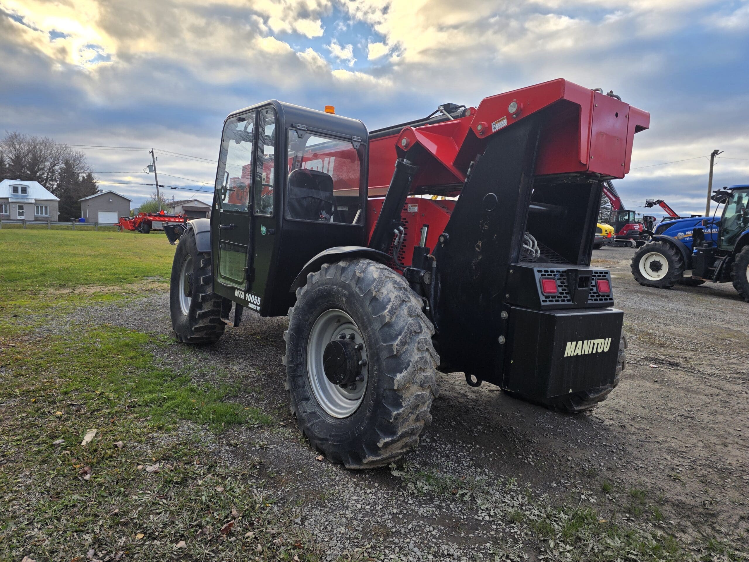 Un télescopique Manitou noir et rouge est garé sur un terrain de gravier et d'herbe sous un ciel nuageux, avec du matériel agricole et des bâtiments visibles à l'arrière-plan.
