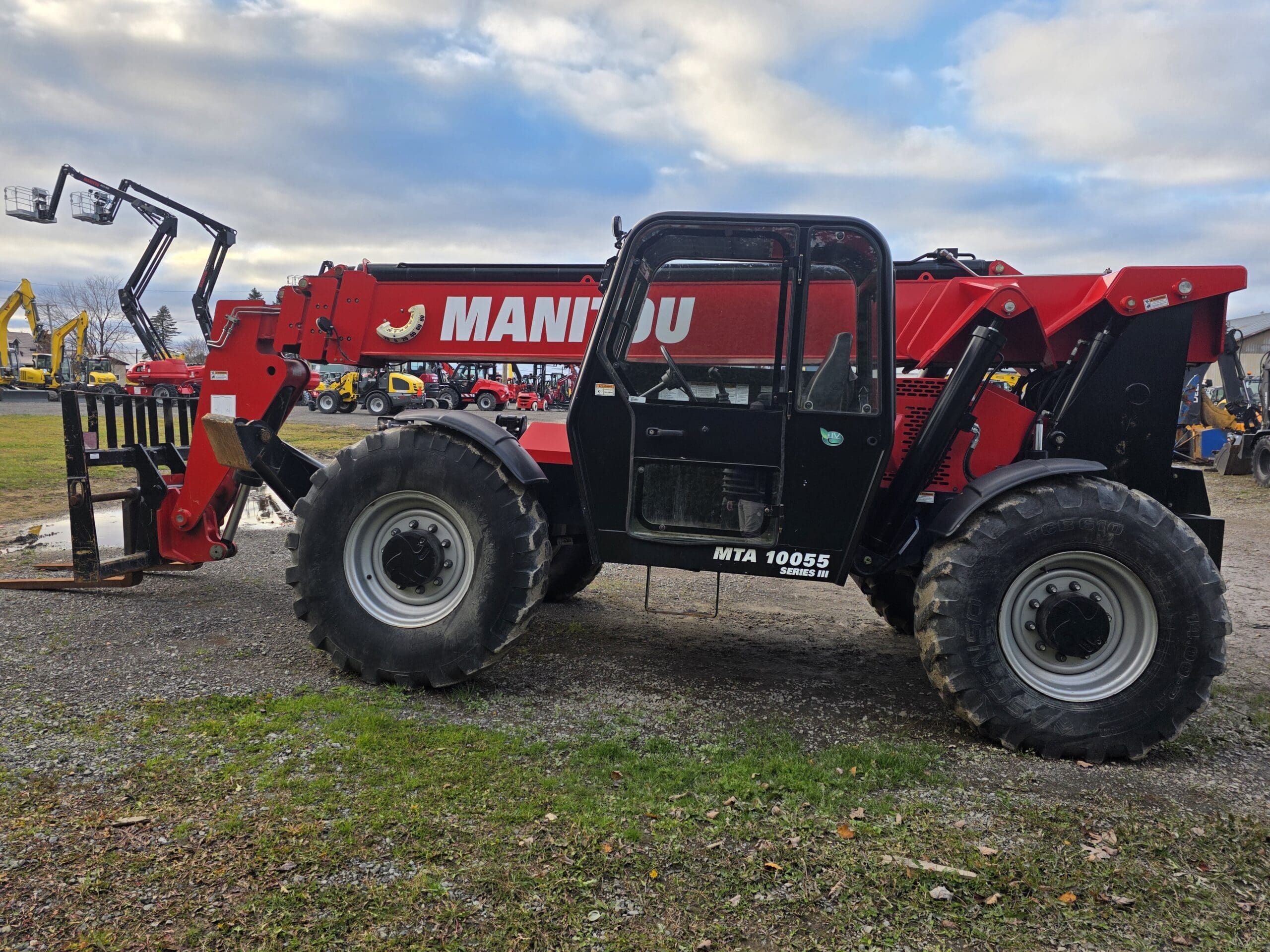 Un télescopique Manitou MTA 10055 rouge et noir avec de gros pneus est garé sur du gravier. La flèche est déployée vers l'avant et des engins de chantier sont visibles à l'arrière-plan sous un ciel partiellement nuageux.