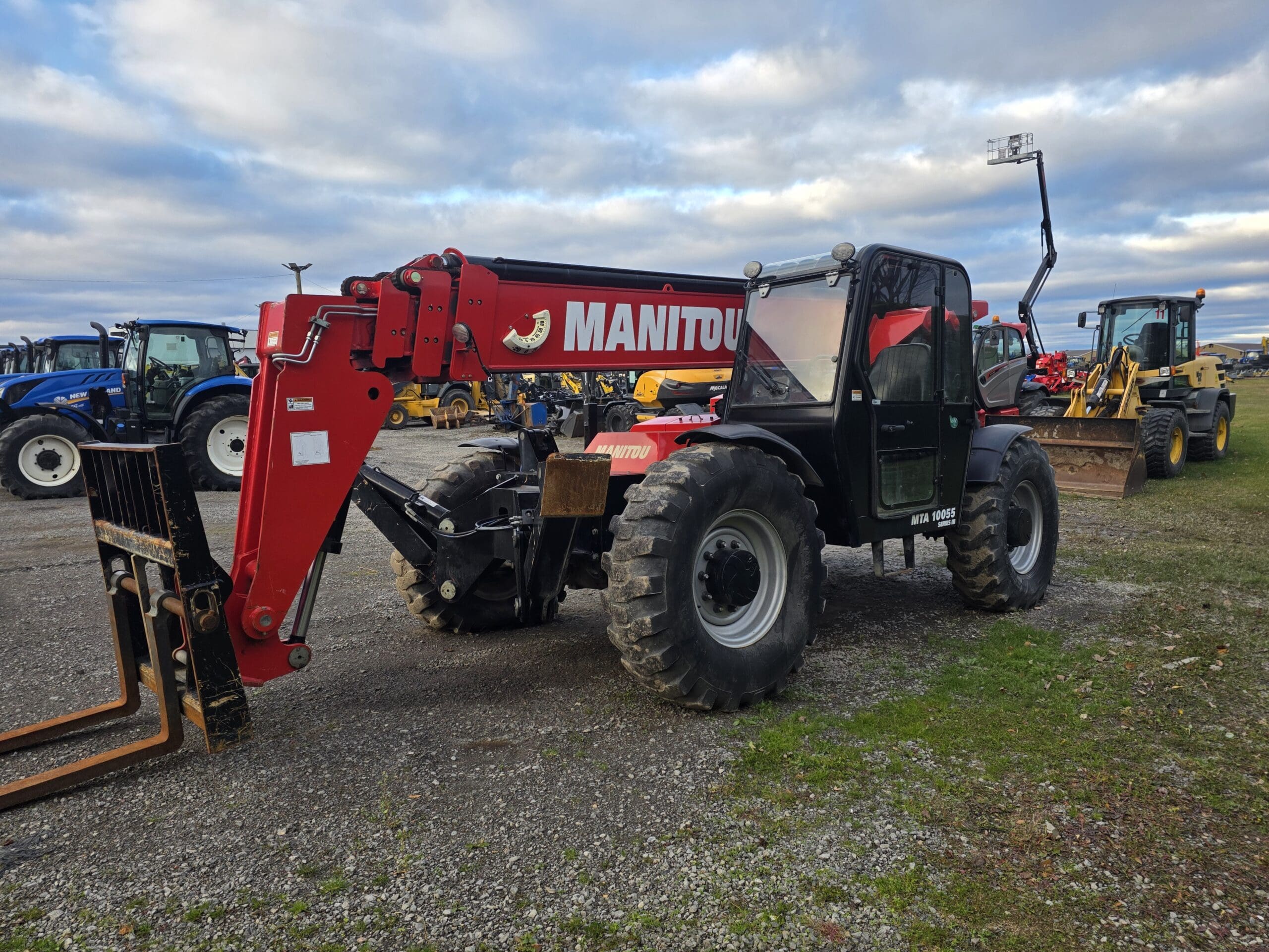 Un télescopique Manitou rouge et noir à gros pneus est garé sur du gravier, entouré d'autres véhicules de construction et agricoles, sous un ciel nuageux.