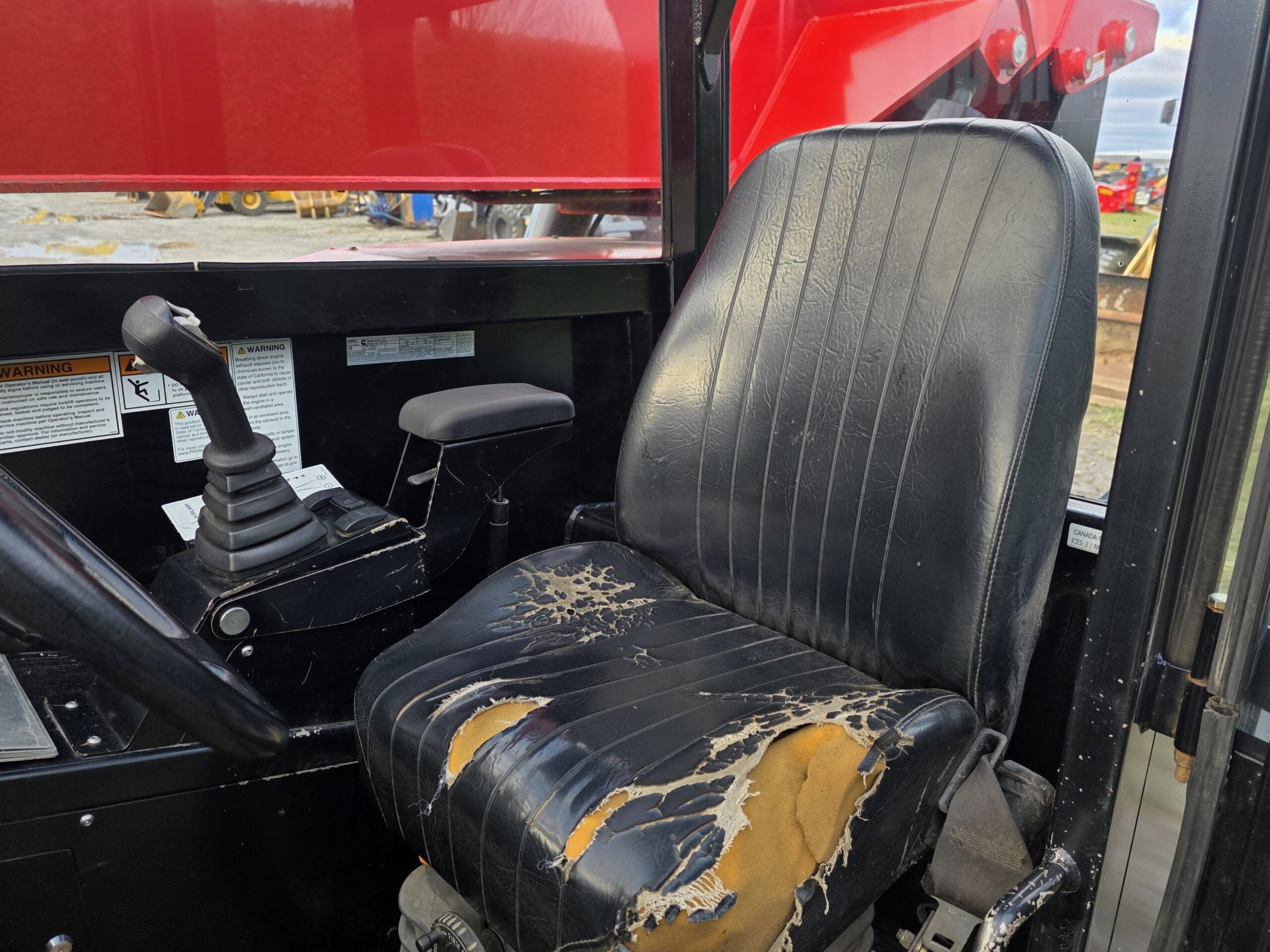 Un siège de conducteur noir, usé et déchiré, avec un rembourrage apparent, à l'intérieur de la cabine d'un véhicule, à côté d'une manette de commande et entouré d'étiquettes d'avertissement ; des machines rouges sont visibles à l'extérieur de la fenêtre.