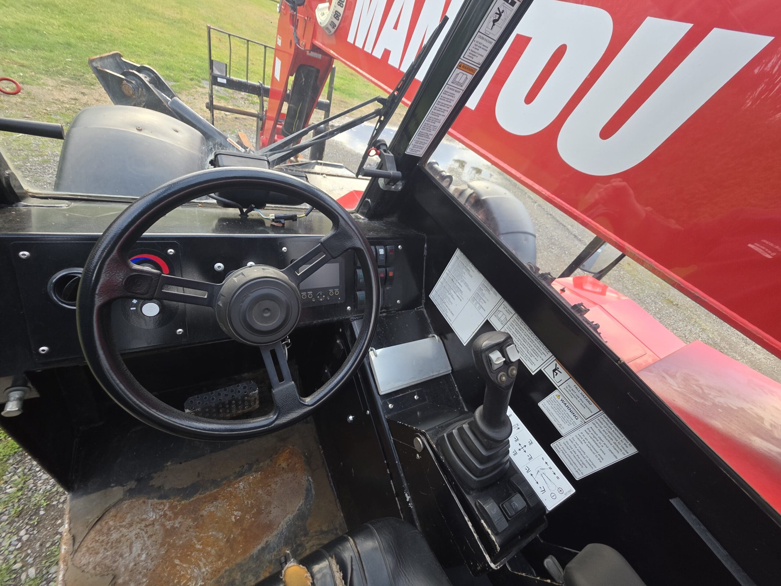 Vue rapprochée de l'intérieur de la cabine d'un véhicule Manitou, montrant le volant, le levier de commande, les pédales, le tableau de bord et diverses étiquettes d'instructions. L'extérieur rouge et la zone herbeuse sont visibles par le côté ouvert.