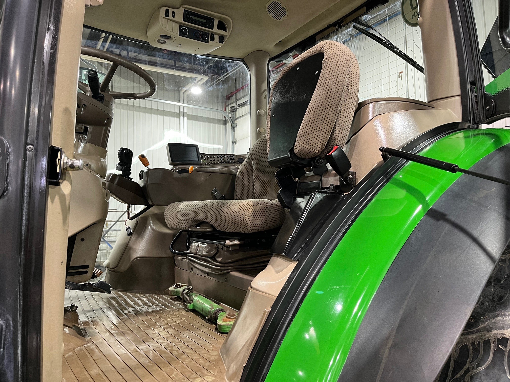 L'image montre l'intérieur d'une cabine de tracteur avec des sièges en tissu, un volant, des leviers de commande et un écran d'affichage. Le sol est recouvert d'un tapis en caoutchouc et la carrosserie verte du tracteur est partiellement visible.