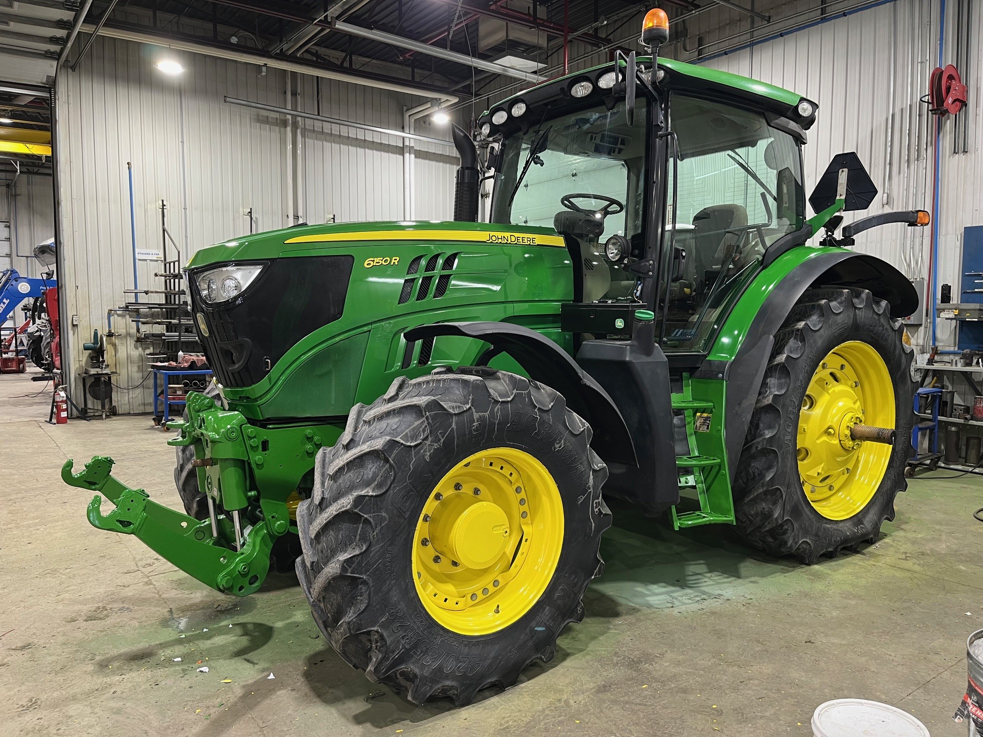 Un tracteur John Deere 6150R vert avec de grandes roues jaunes est garé à l'intérieur d'un garage ou d'un atelier spacieux et bien éclairé, avec des sols en béton et divers outils en arrière-plan.