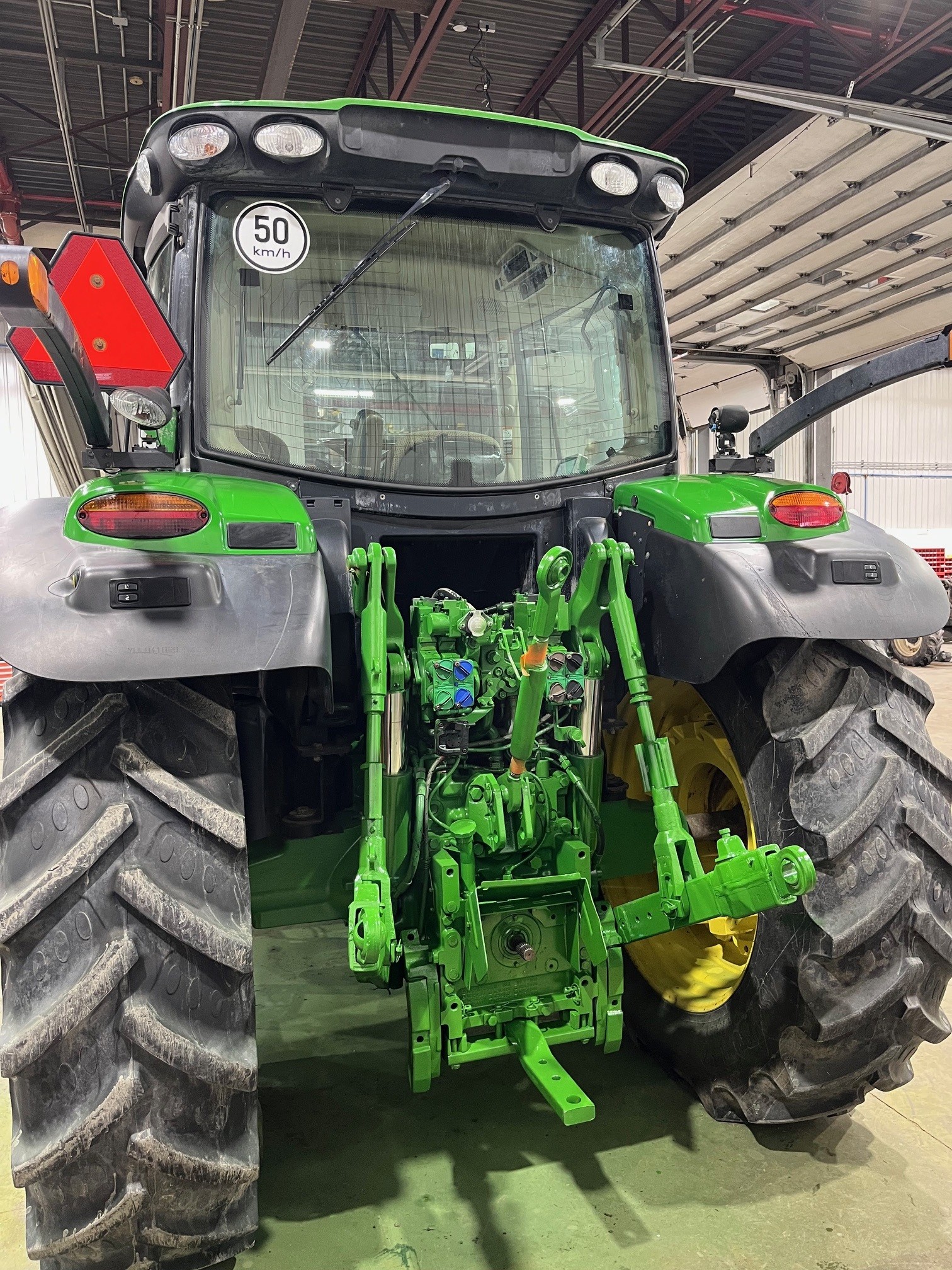 Vue arrière d'un tracteur vert garé à l'intérieur, montrant de gros pneus et des équipements hydrauliques. Le tracteur se trouve dans un garage ou un atelier bien éclairé, avec des outils et des équipements visibles à l'arrière-plan.
