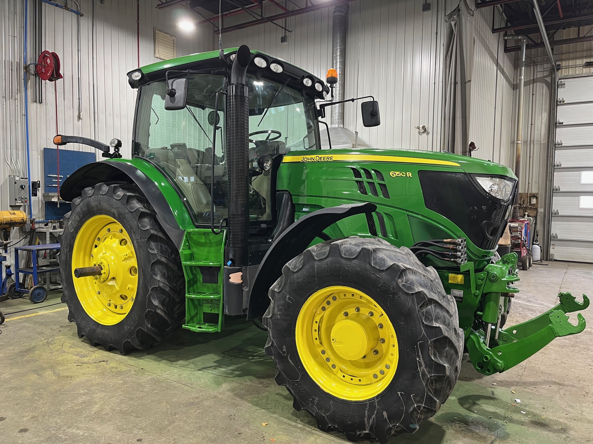 Un tracteur John Deere 6155R vert et jaune est garé à l'intérieur d'un grand atelier bien éclairé, avec des sols en béton et des murs en métal. Divers outils et équipements sont visibles à l'arrière-plan.