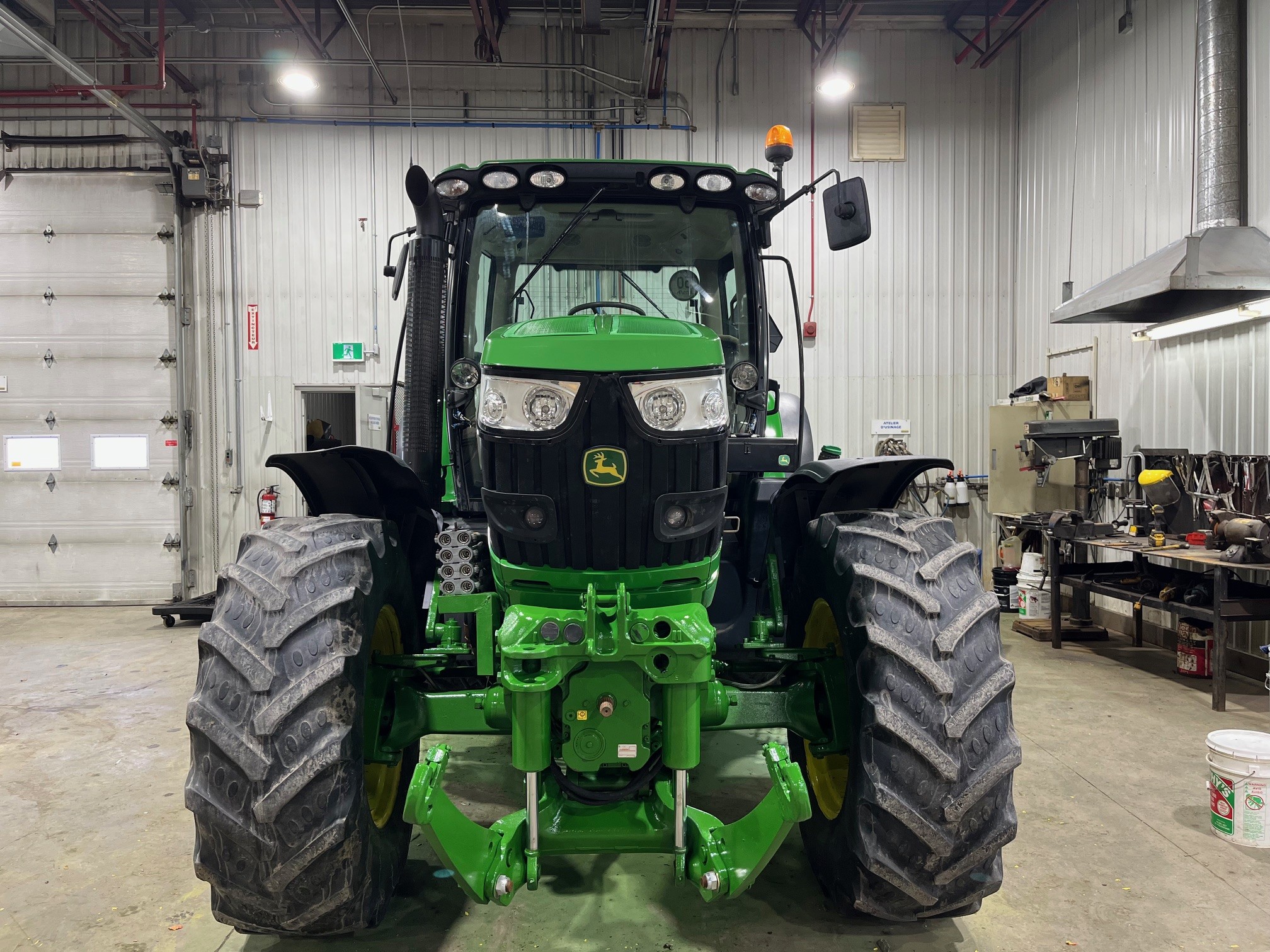 Un tracteur John Deere vert est garé à l'intérieur d'un atelier industriel spacieux avec des sols en béton, des murs en métal et divers outils et équipements le long du mur latéral.