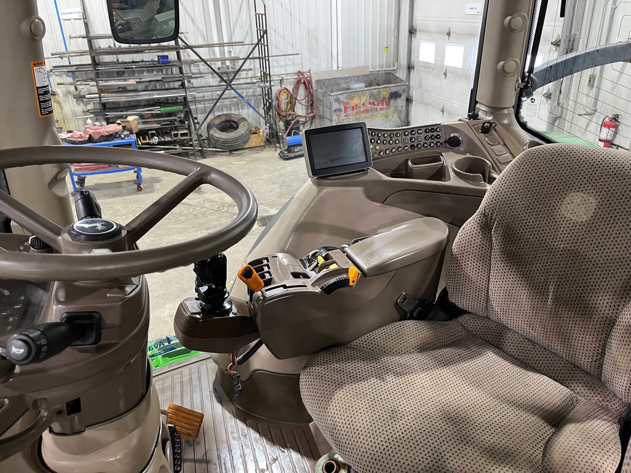 Intérieur d'une cabine de tracteur agricole avec un siège rembourré, un panneau de commande, un joystick et un écran d'affichage, à l'intérieur d'un atelier avec des outils, des pneus, des échafaudages et des équipements visibles à travers la fenêtre de la cabine.