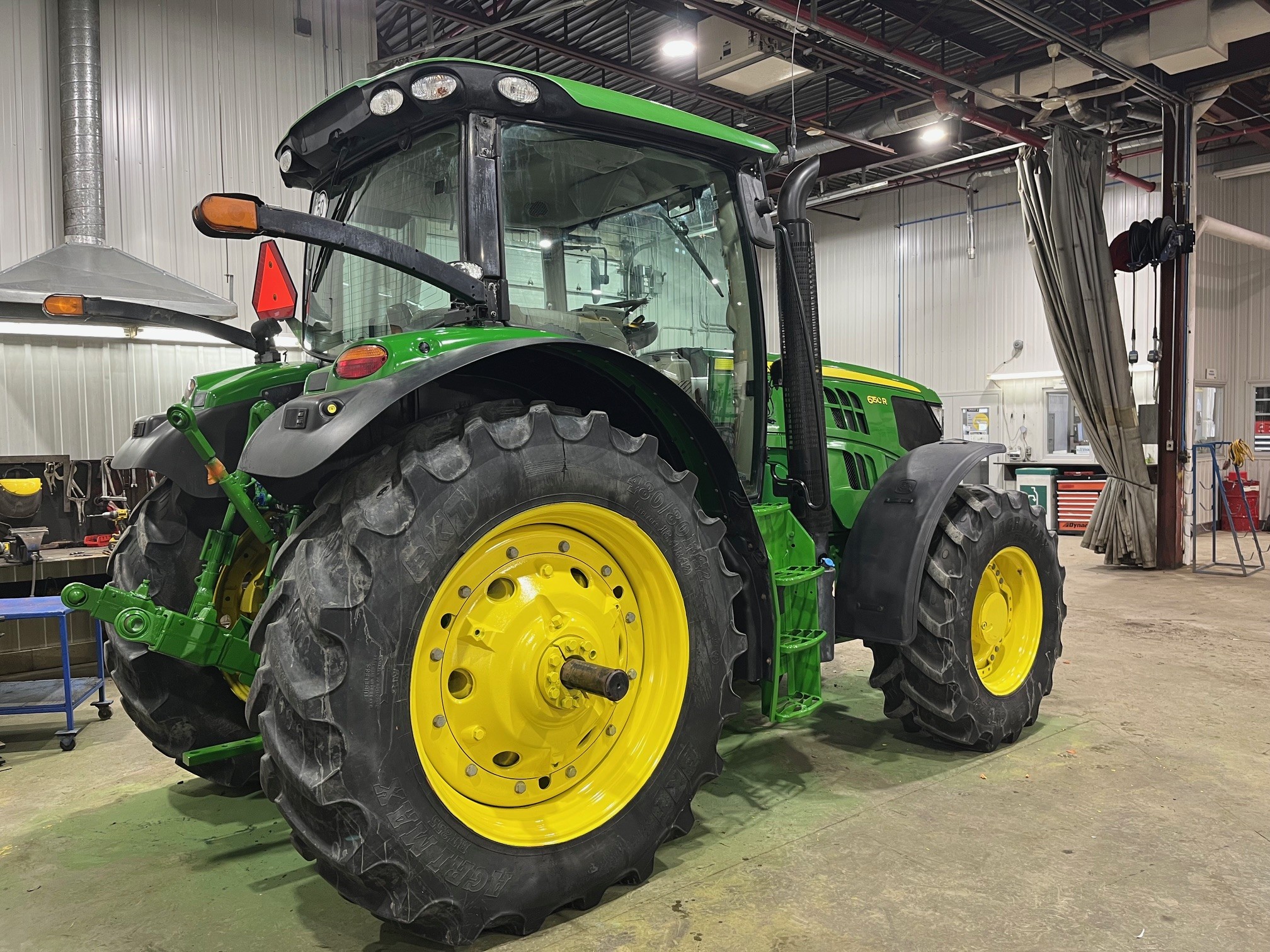 Un tracteur John Deere vert et jaune est garé à l'intérieur d'un atelier bien éclairé, avec des sols en béton et des outils et équipements visibles à l'arrière-plan.