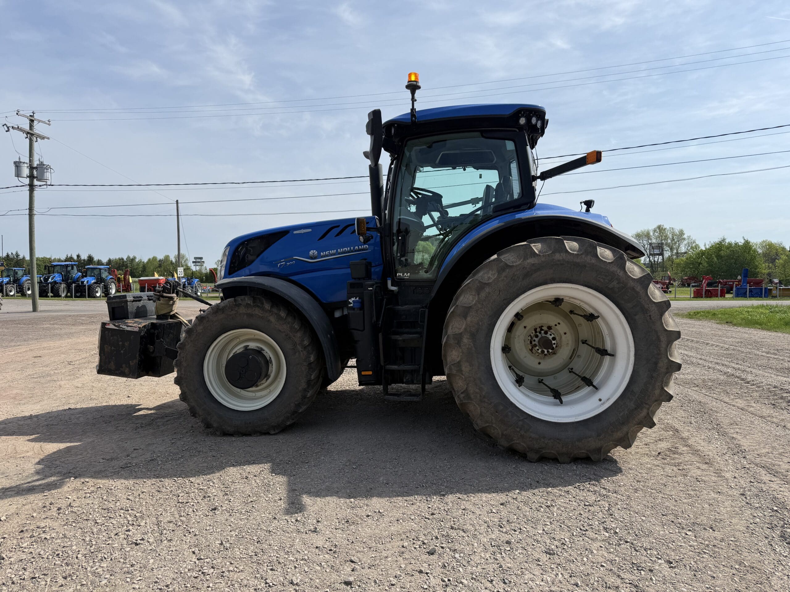 Un tracteur New Holland bleu à gros pneus est garé sur une surface de gravier sous un ciel partiellement nuageux, avec des terres agricoles et des poteaux électriques visibles à l'arrière-plan.