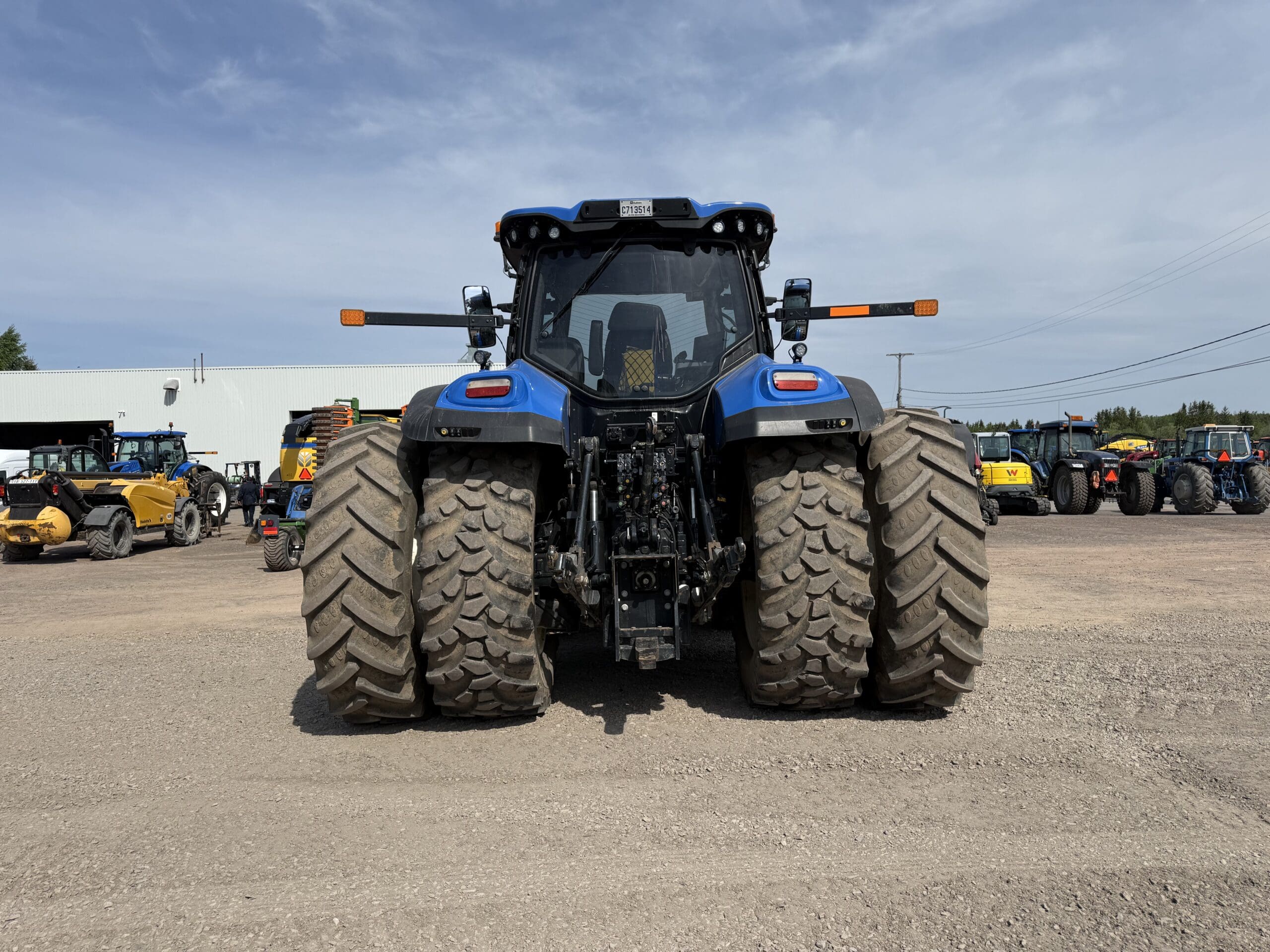 Un grand tracteur bleu avec des pneus arrière jumelés est garé sur un terrain en gravier, face à la caméra. D'autres tracteurs et machines sont visibles à l'arrière-plan, près d'un bâtiment blanc, sous un ciel partiellement nuageux.