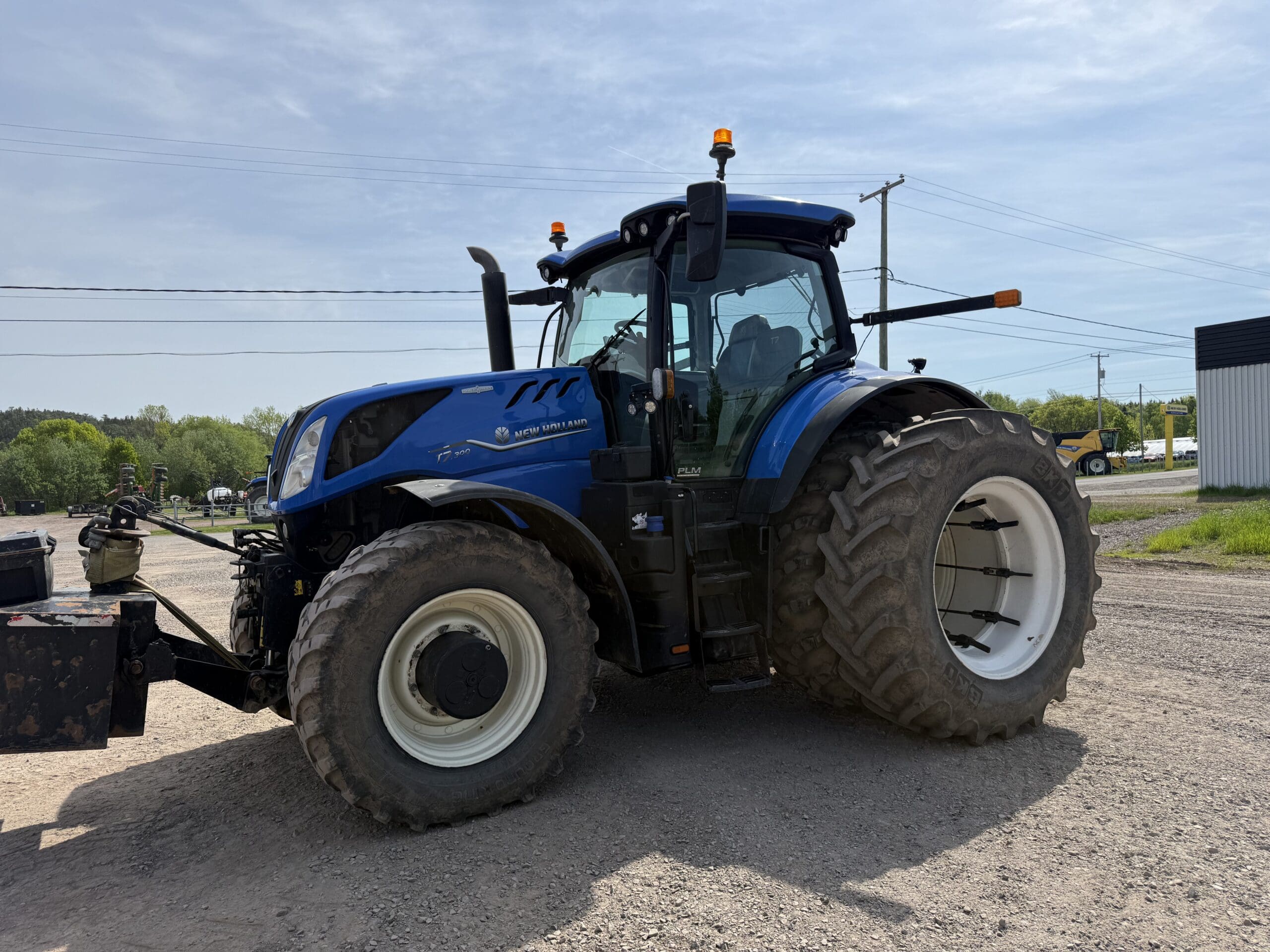 Un grand tracteur bleu aux pneus arrière surdimensionnés est garé sur une route de gravier sous un ciel clair, avec des arbres, des lignes électriques et un bâtiment visibles à l'arrière-plan.