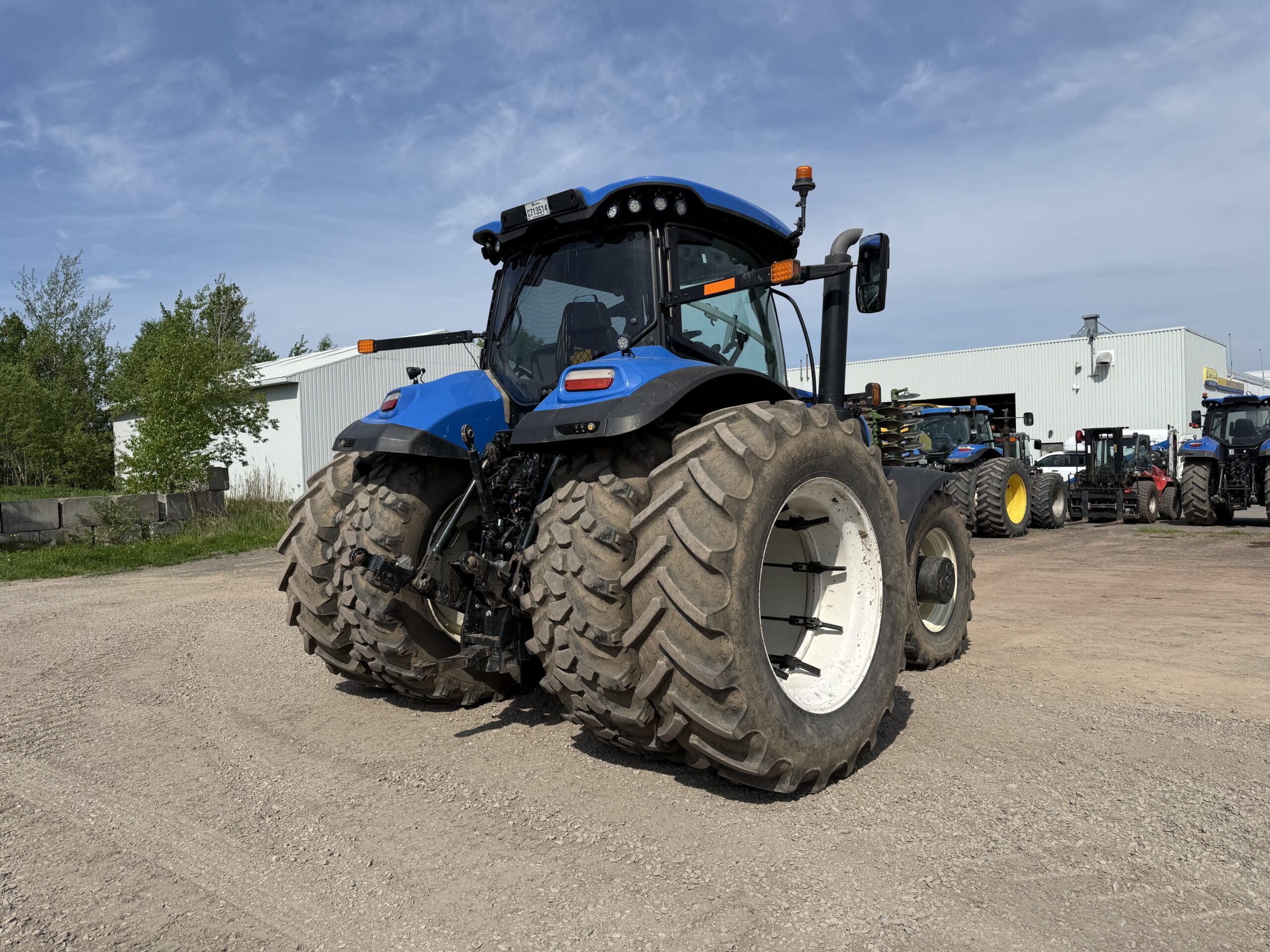 Un grand tracteur bleu à roues arrière jumelées est garé sur un terrain en gravier près de bâtiments industriels blancs et d'autres tracteurs, sous un ciel partiellement nuageux.