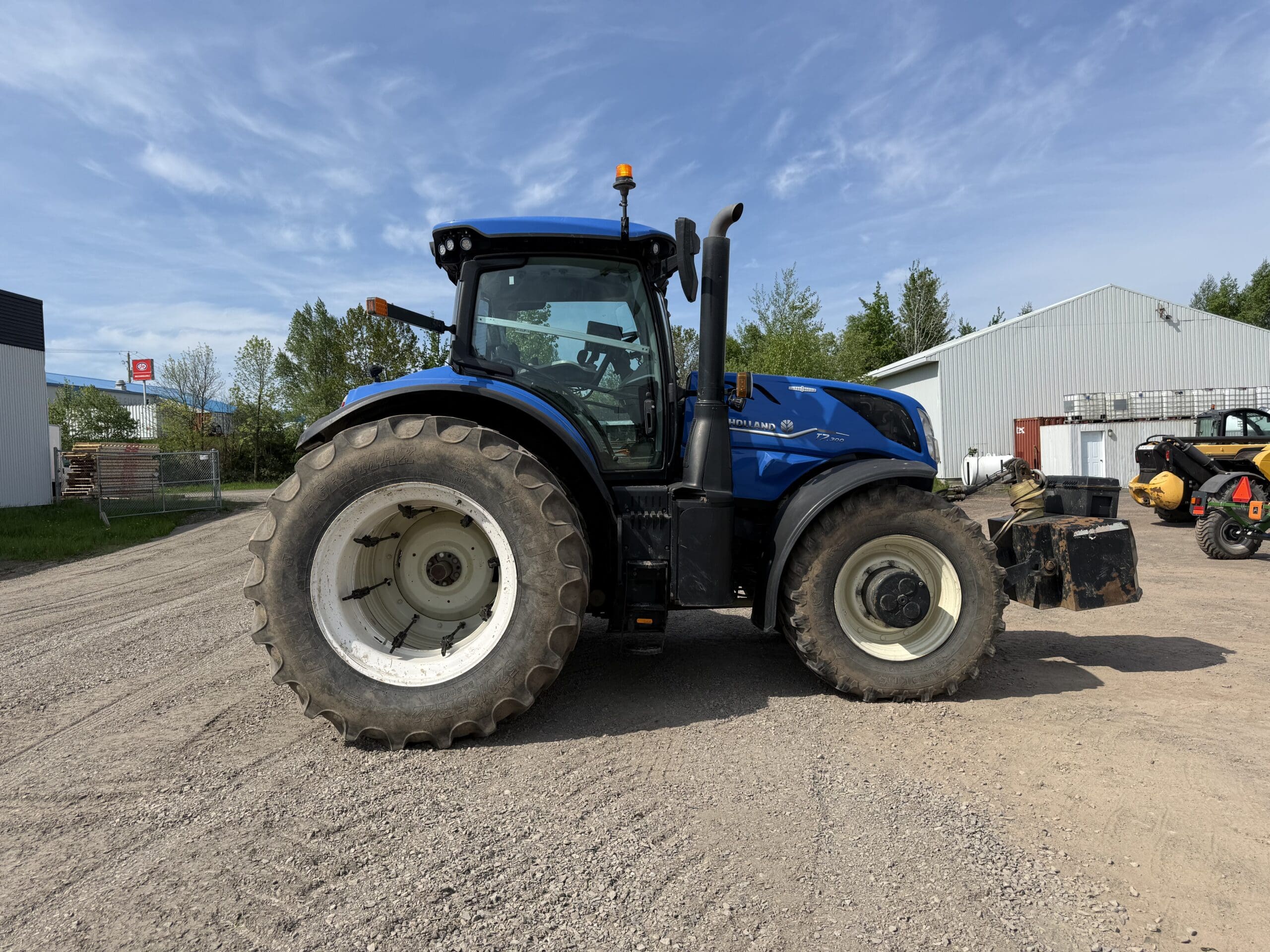 Un tracteur bleu à gros pneus est garé sur une surface de gravier près de bâtiments industriels et de quelques arbres, sous un ciel bleu avec des nuages épars.