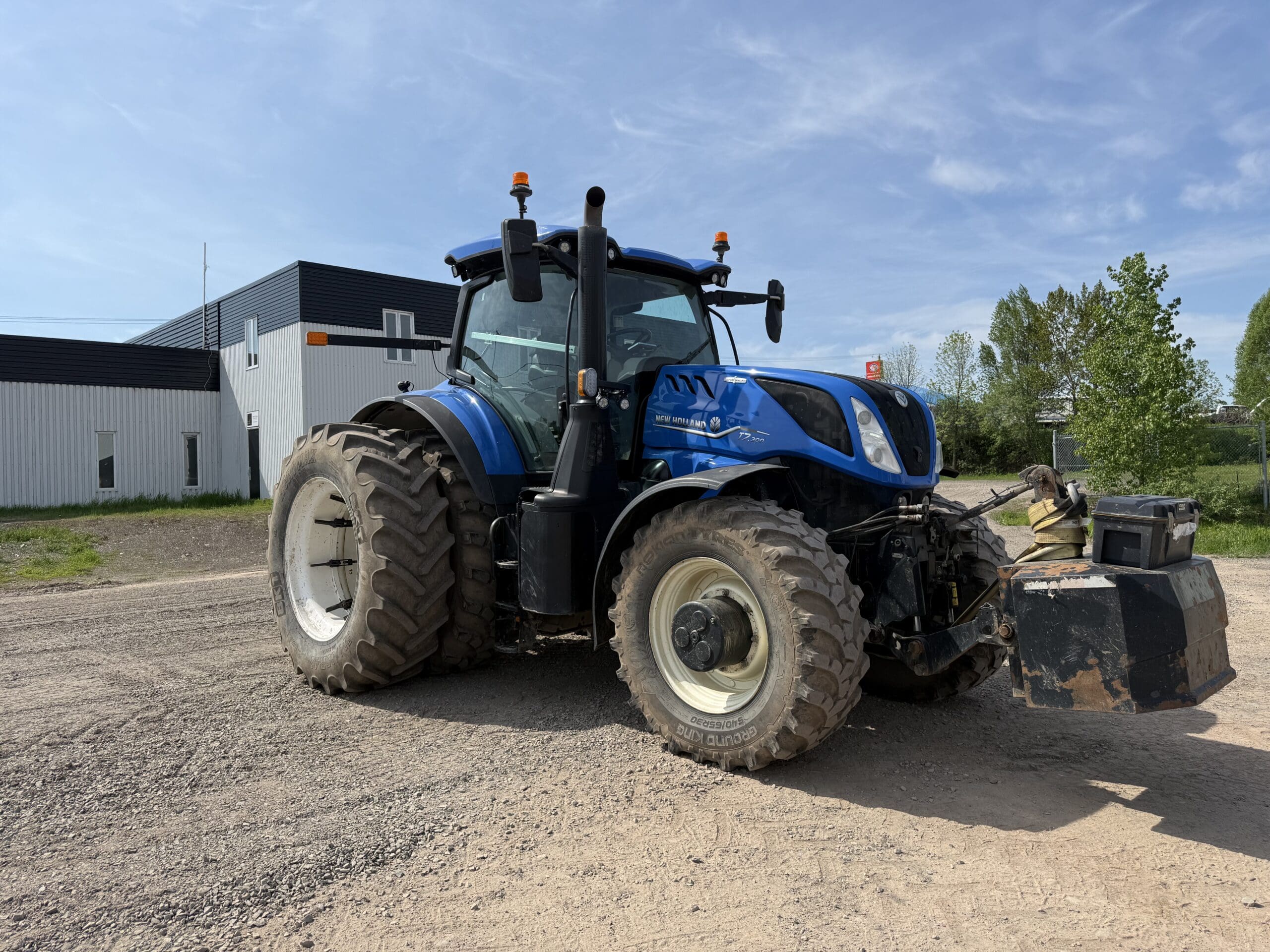 Un grand tracteur bleu à pneus larges est garé sur une surface de gravier près d'un bâtiment industriel blanc, avec des arbres verts et un ciel bleu en arrière-plan.