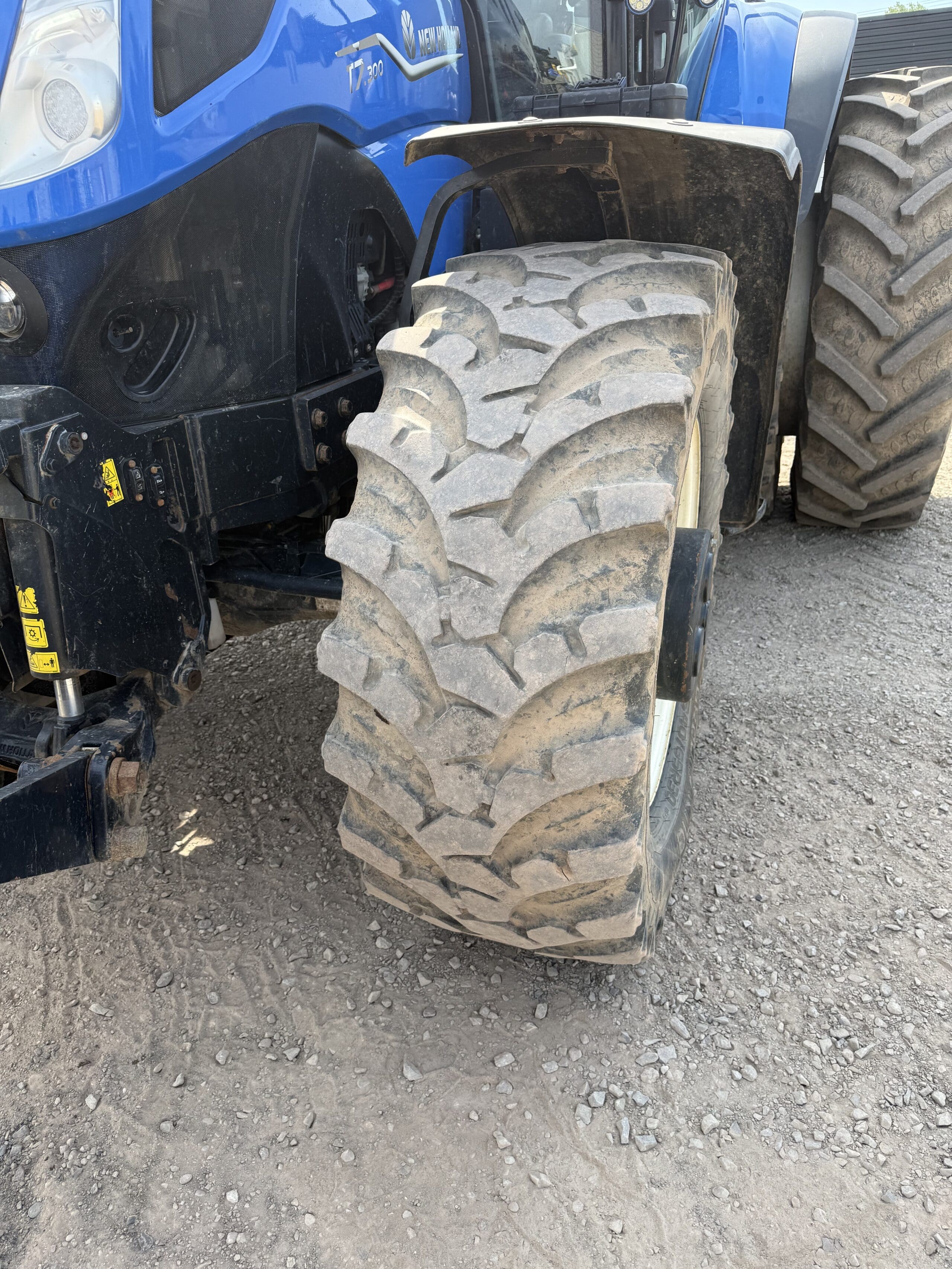 Gros plan d'un gros pneu de tracteur robuste attaché à un tracteur bleu, garé sur un sol recouvert de gravier, avec un autre pneu visible à l'arrière-plan.