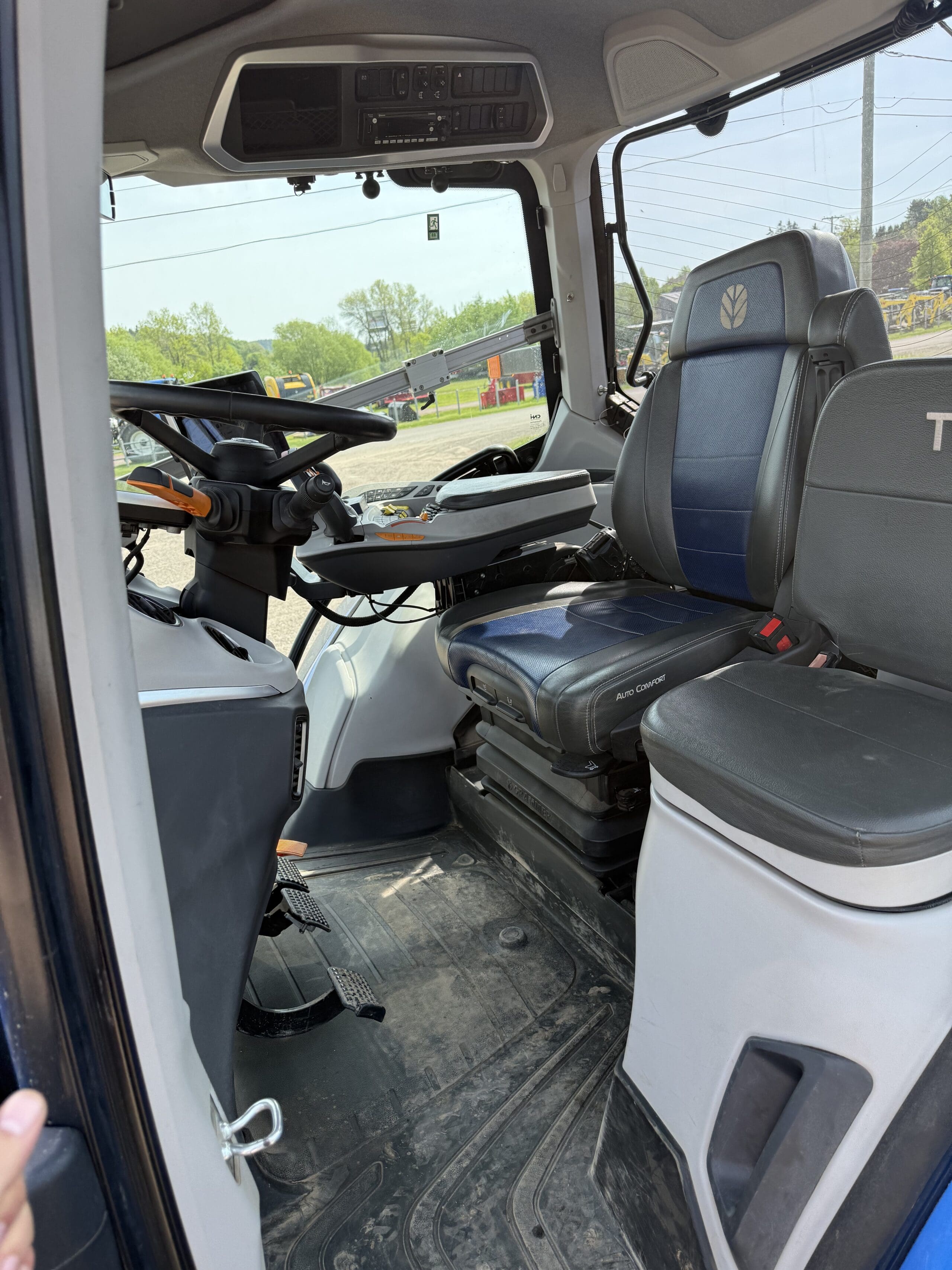 L'intérieur d'une cabine de tracteur avec un volant, un panneau de commande, des sièges conducteur et passager bleu foncé et gris, et de grandes fenêtres montrant un chantier de construction à l'extérieur.