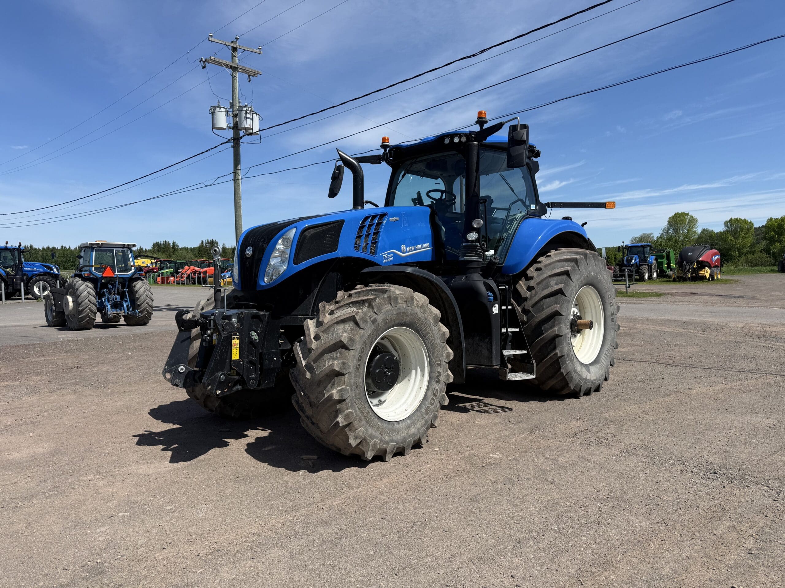 Un grand tracteur bleu est garé sur un terrain en terre battue, sous un ciel d'un bleu limpide, tandis qu'on aperçoit d'autres tracteurs et du matériel agricole à l'arrière-plan.